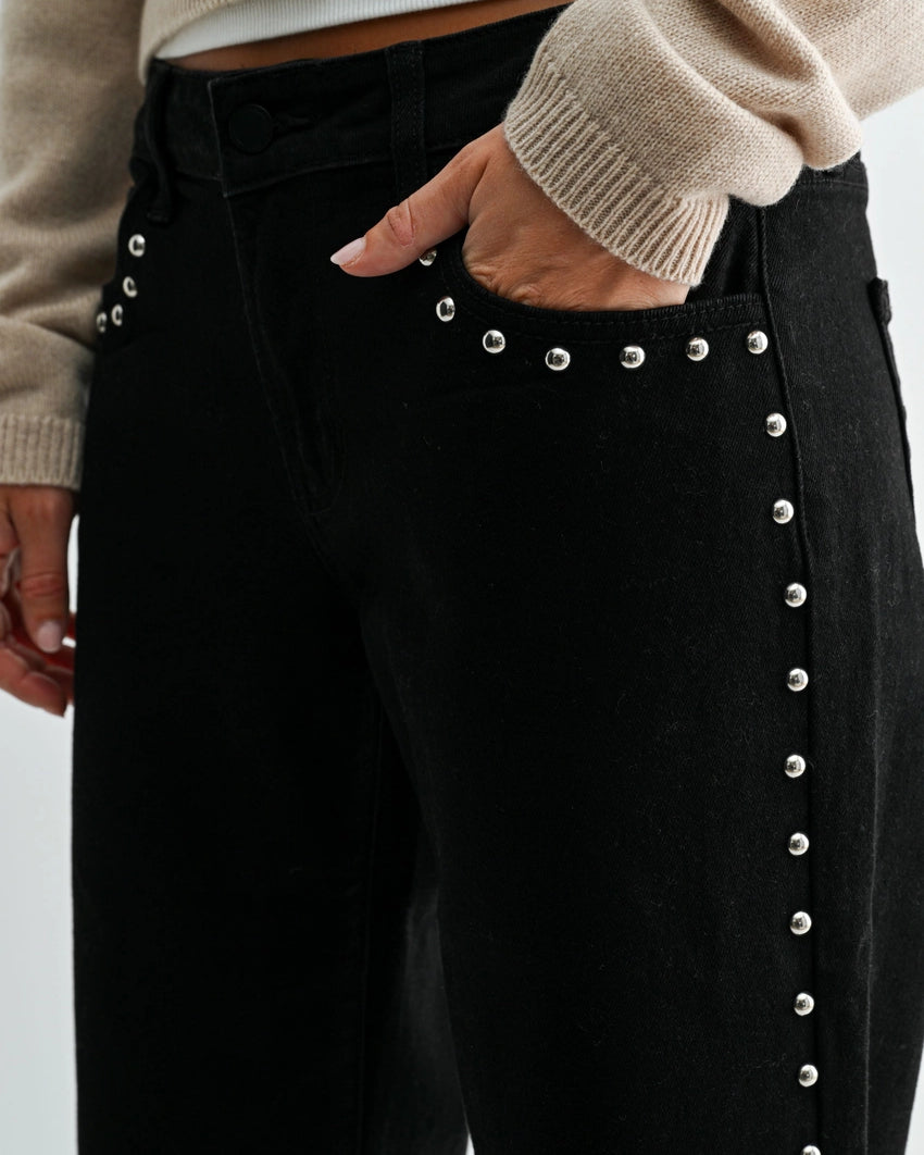 woman wearing black jeans with silver studs and a cream sweater in front of a white background with her hand in the front pocket.