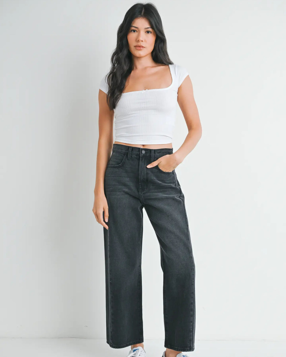 woman wearing black jeans and white top in front of a white background.