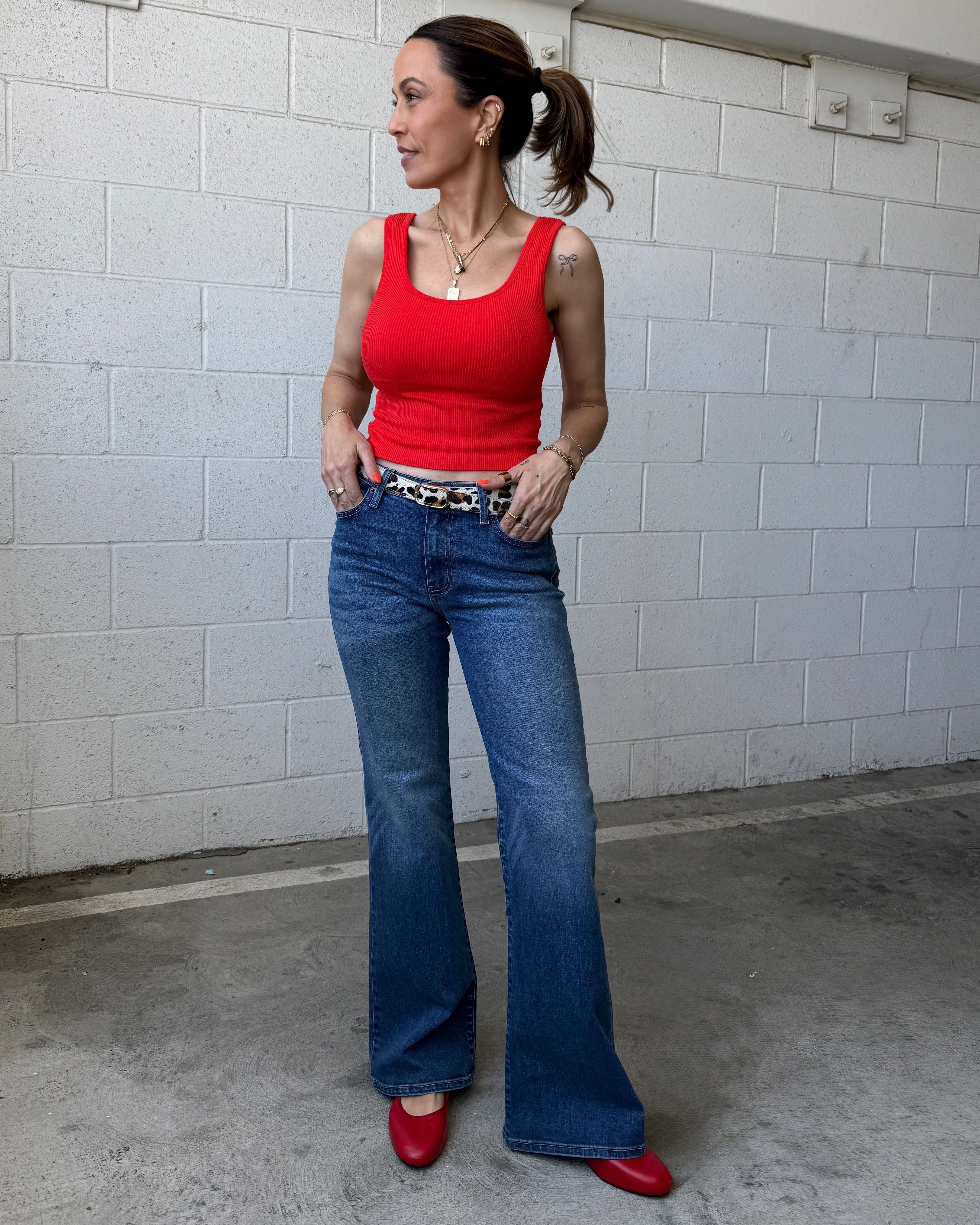 Woman wearing a red tank top and blue jeans standing against a white tiled wall.