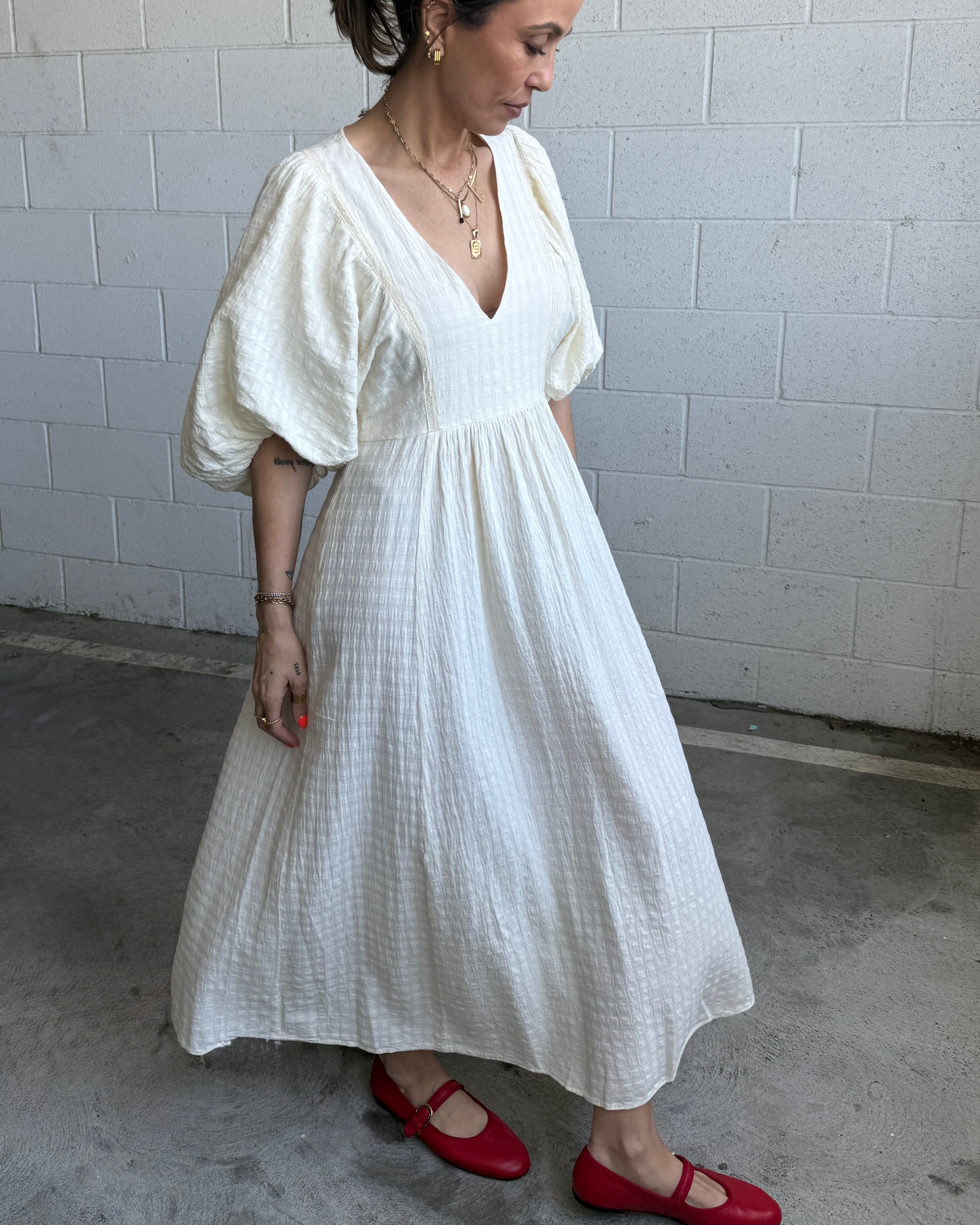 Woman wearing a white dress with puffed sleeves and red shoes against a gray brick wall.