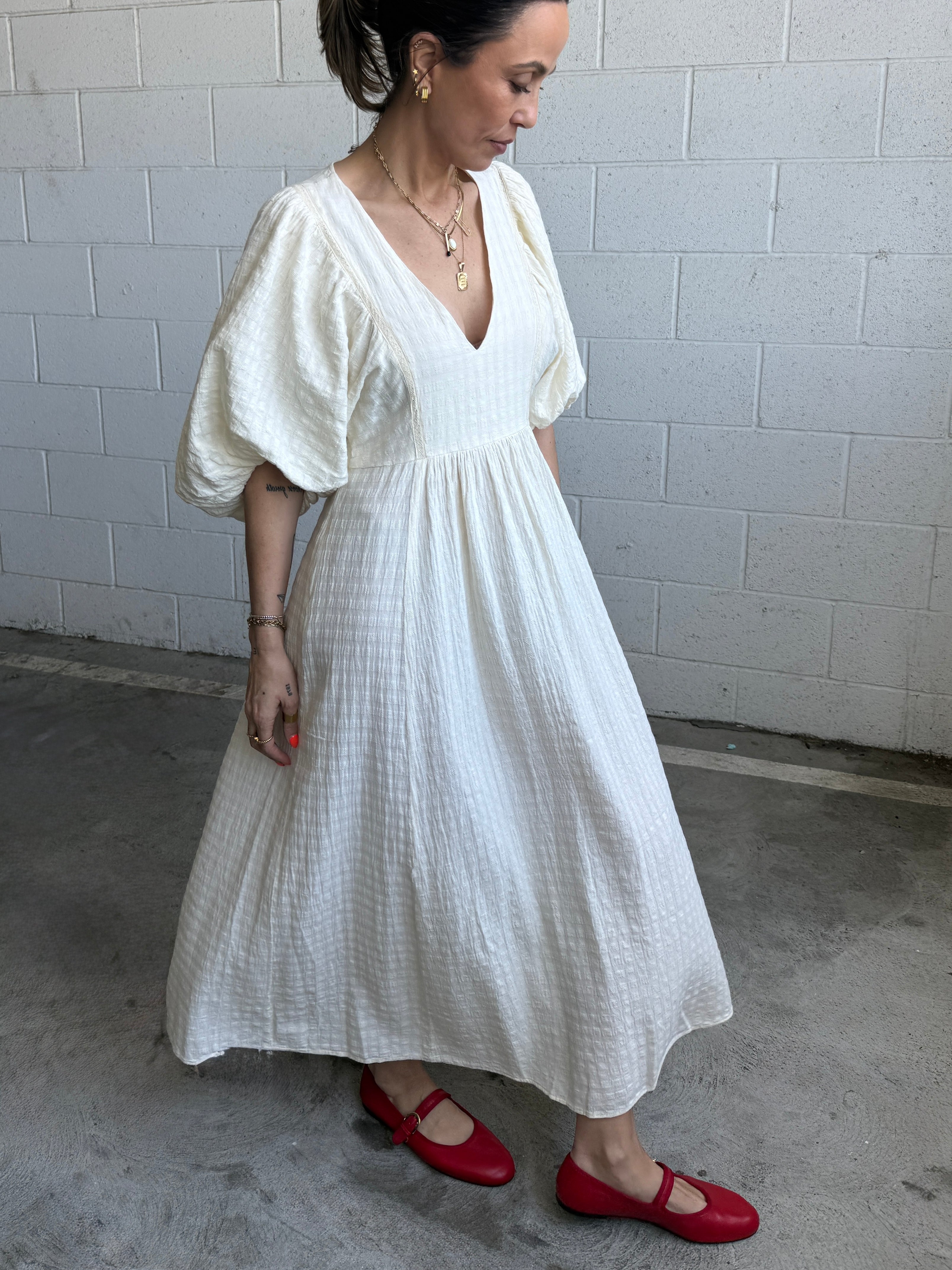 Woman wearing a white dress with puffed sleeves and red shoes against a gray brick wall.