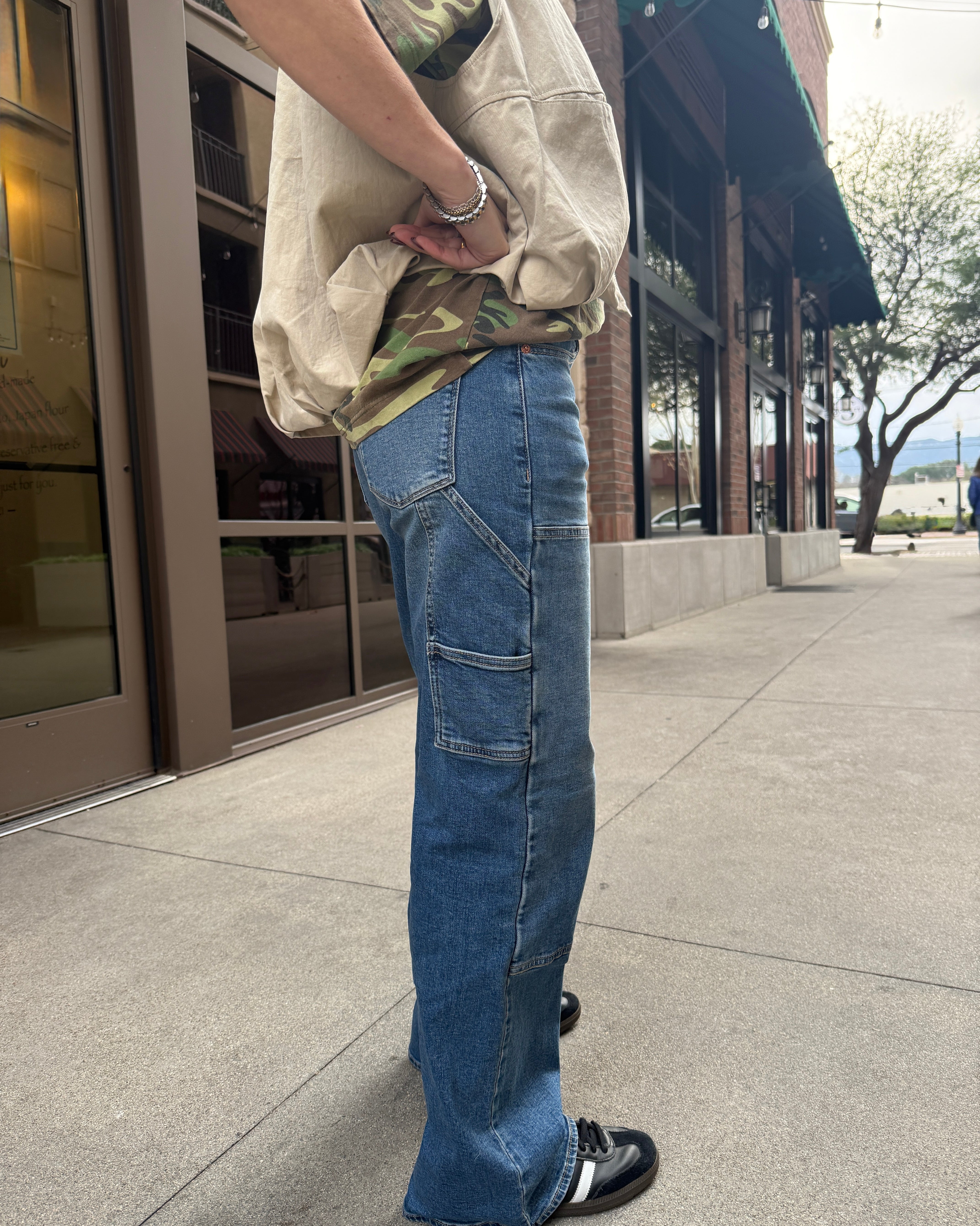 Woman wearing carpenter jeans in urban setting with tan vest and camo t-shirt