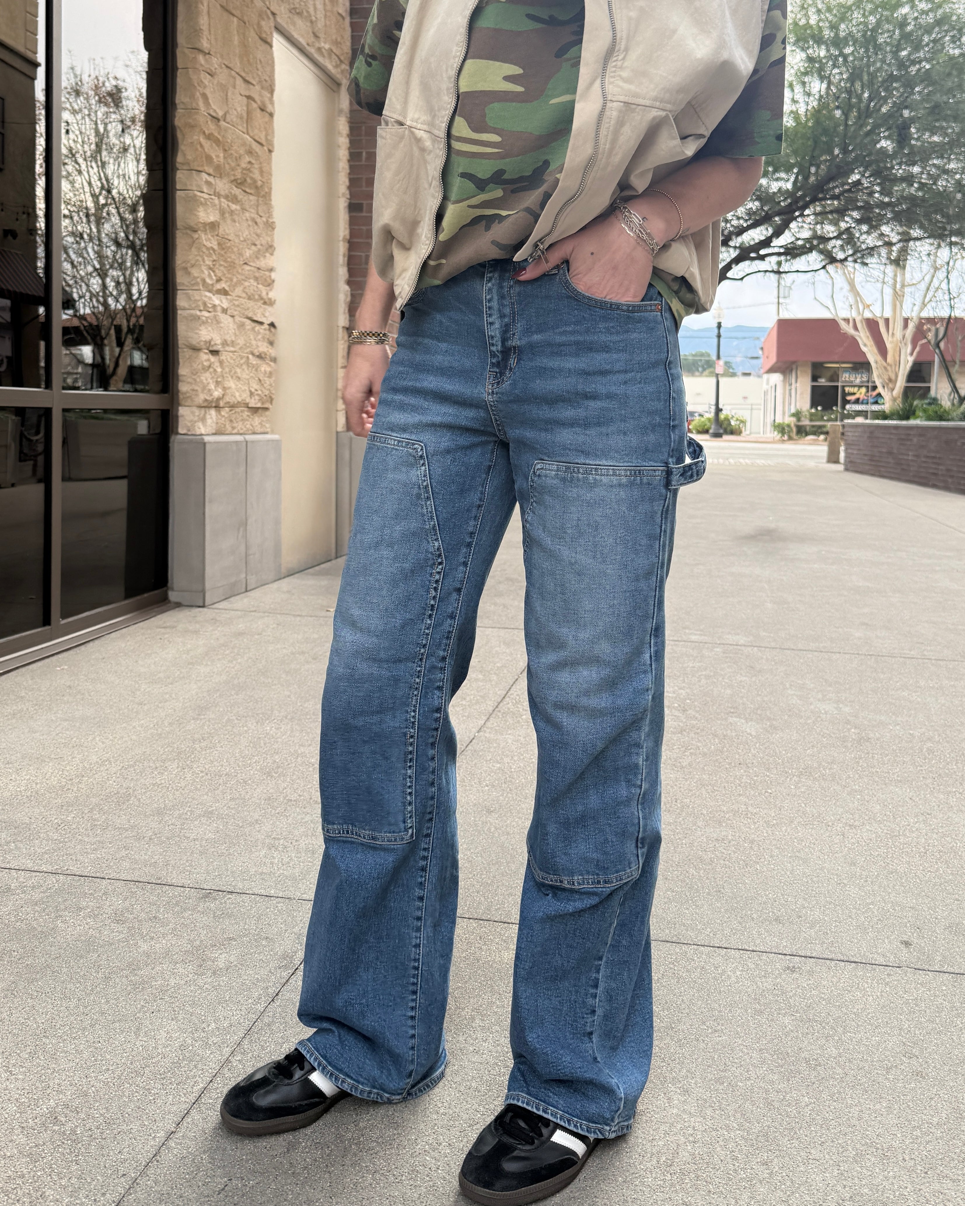 Woman wearing carpenter jeans in urban setting with tan vest and camo t-shirt