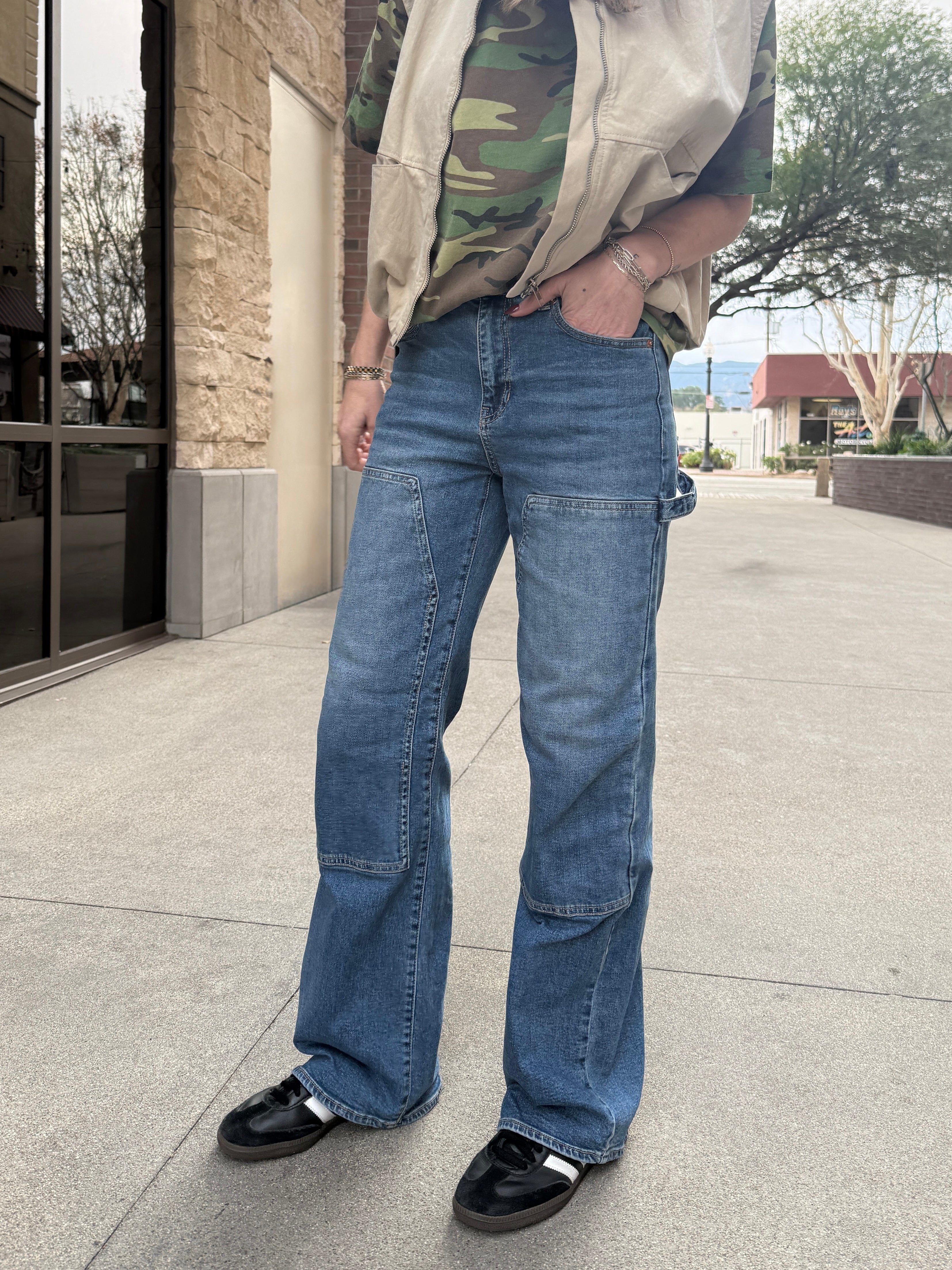 Woman wearing carpenter jeans in urban setting with tan vest and camo t-shirt