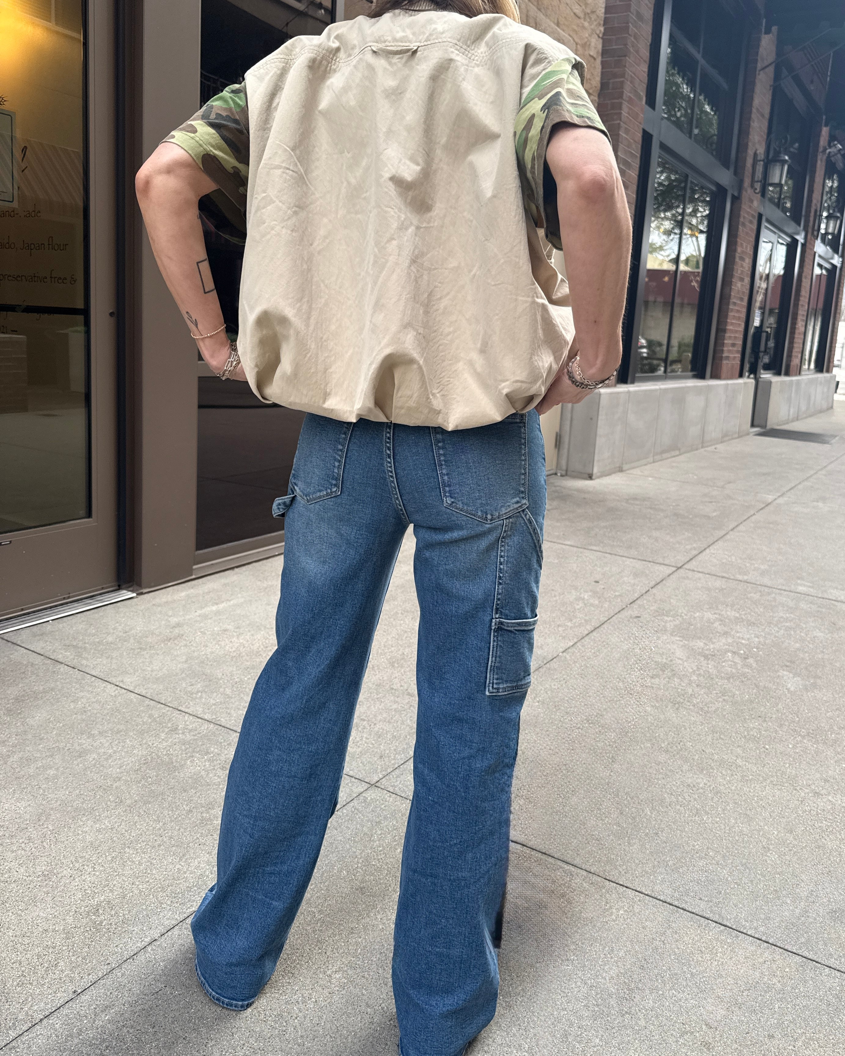 Woman wearing carpenter jeans in urban setting with tan vest and camo t-shirt