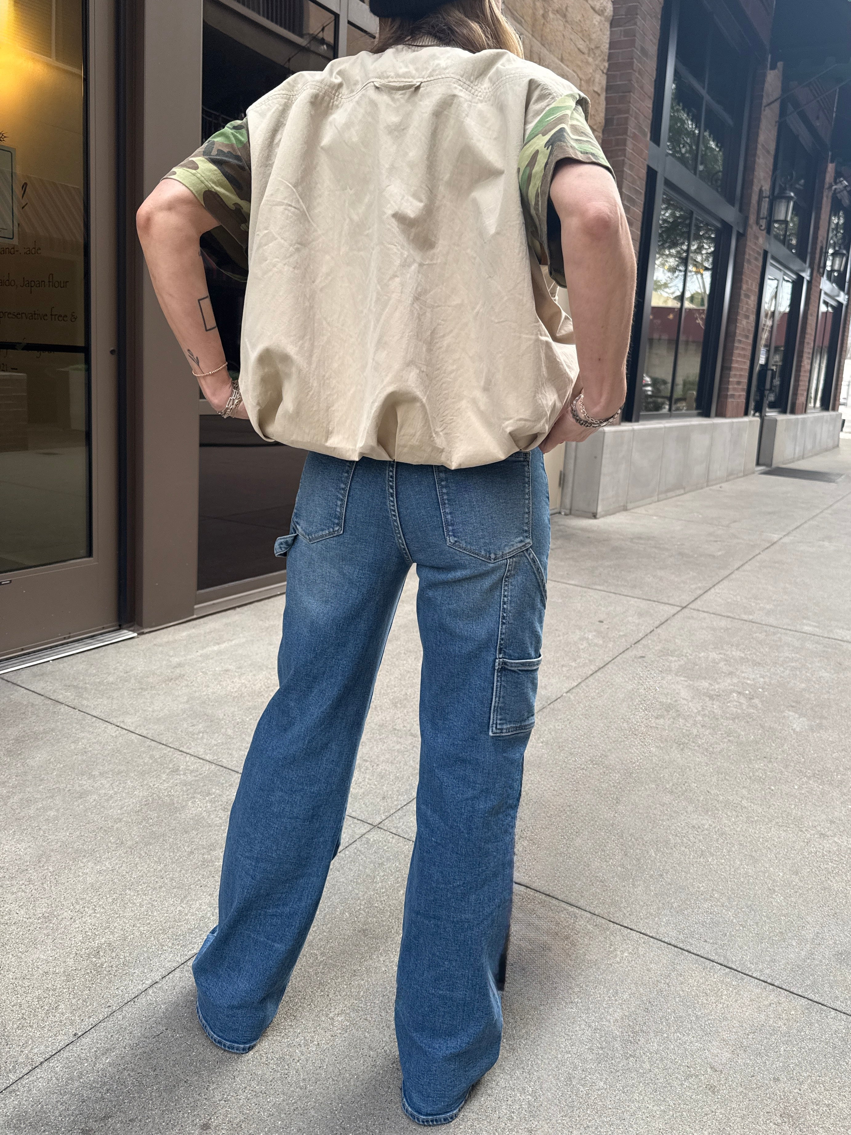Woman wearing carpenter jeans in urban setting with tan vest and camo t-shirt