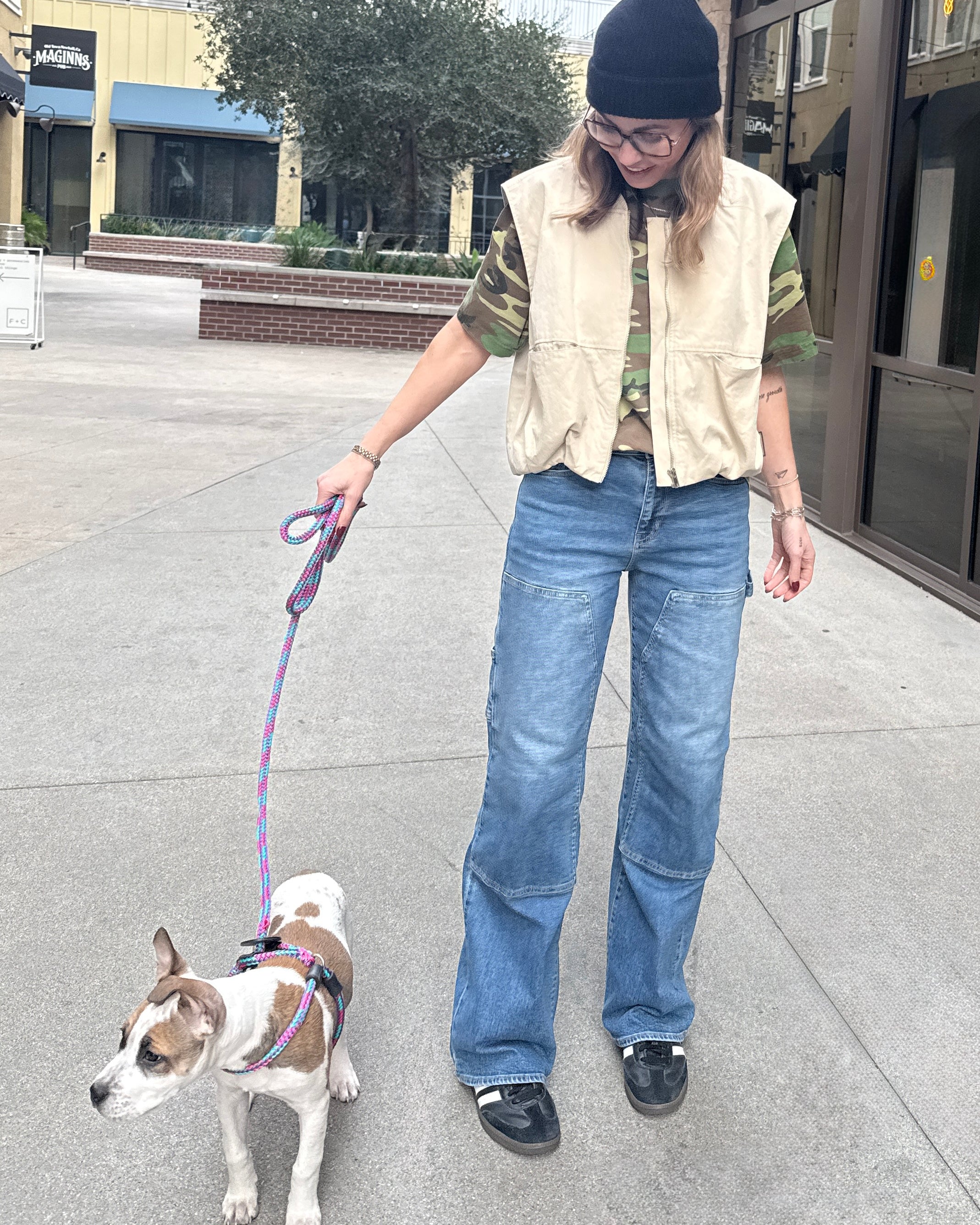 Woman wearing carpenter jeans in urban setting with tan vest and camo t-shirt walking a dog