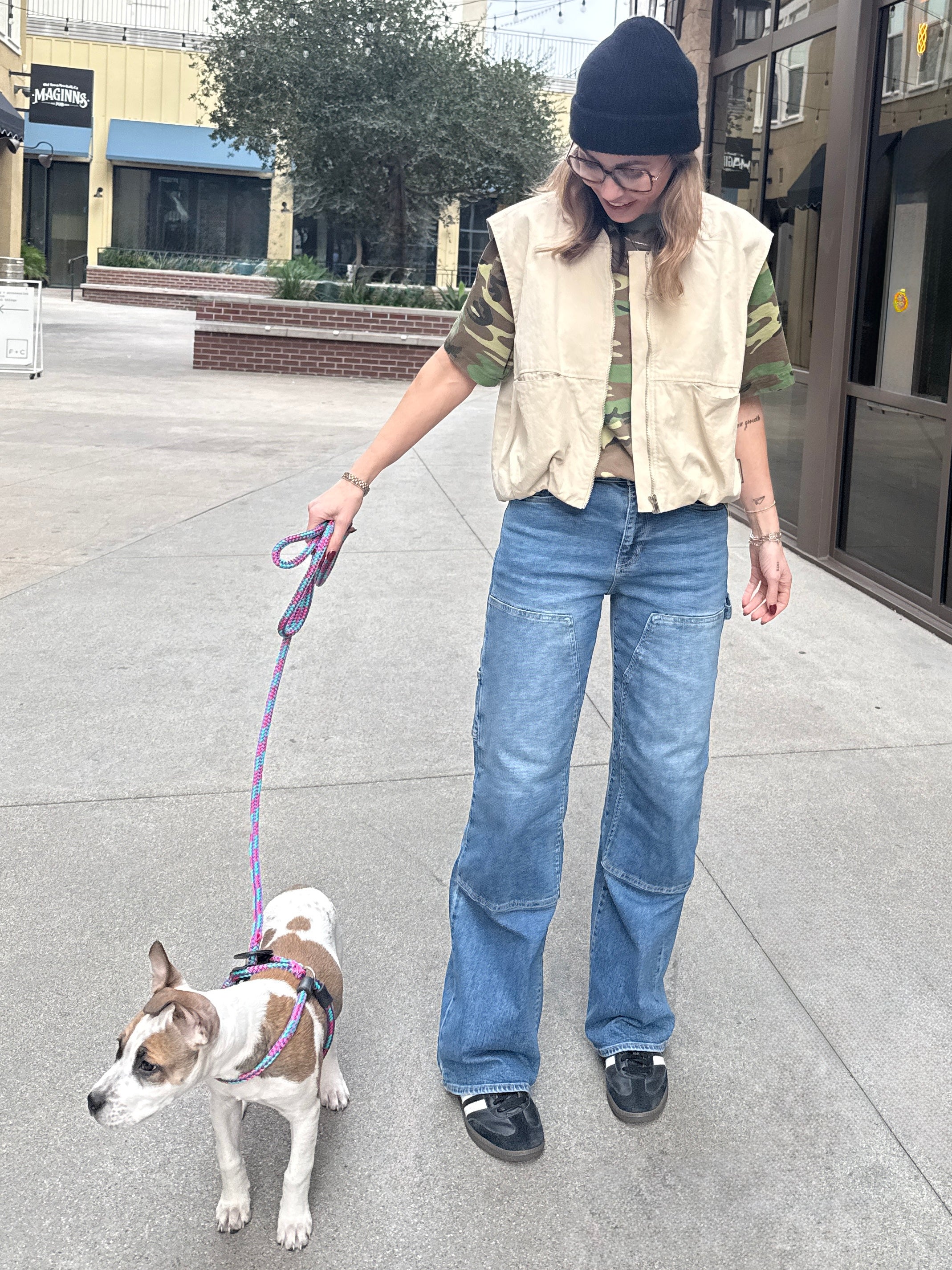 Woman wearing carpenter jeans in urban setting with tan vest and camo t-shirt walking a dog