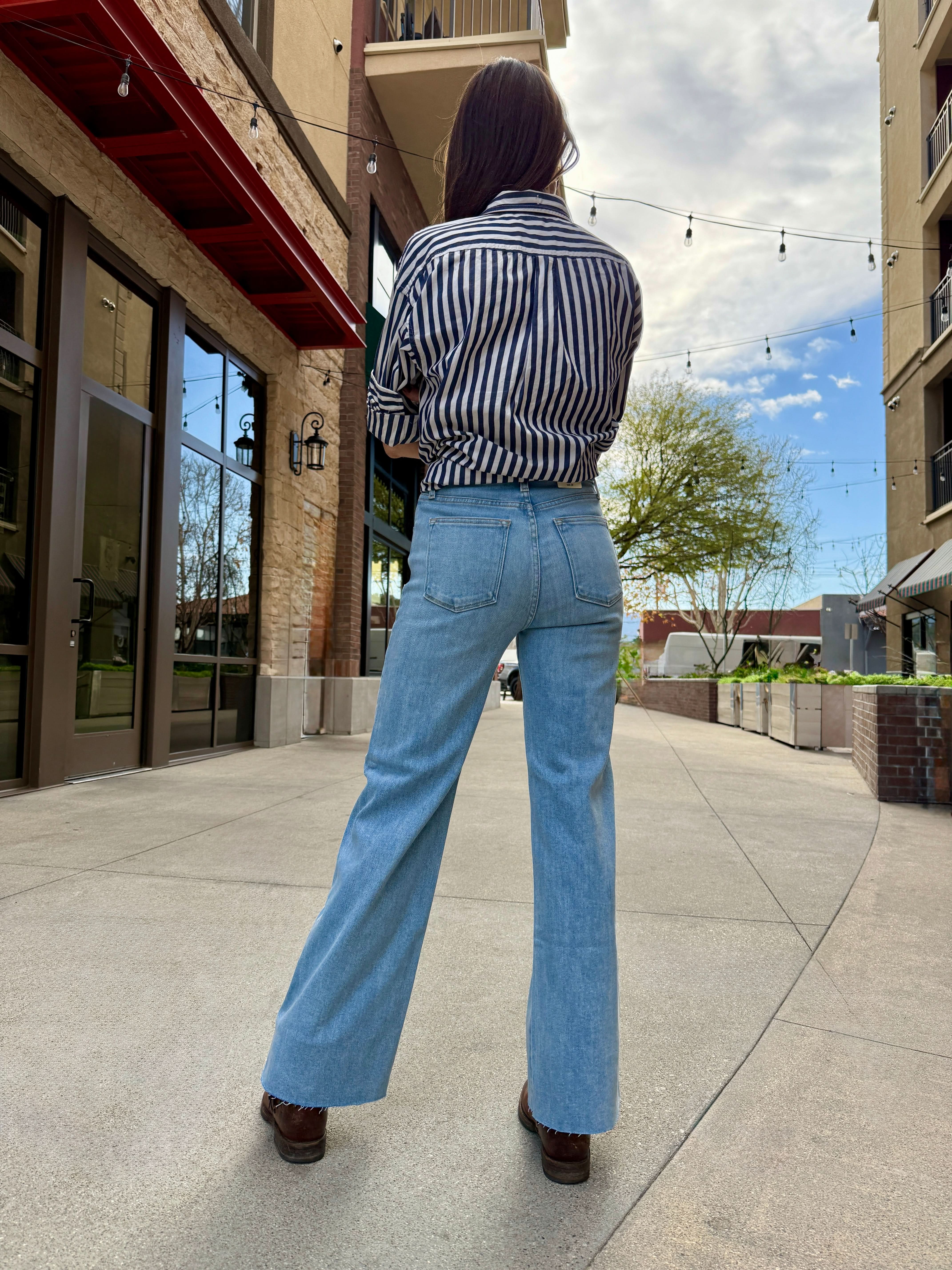 Woman standing outside wearing light wash raw hem jeans with navy and white stripe blouse facing away from the camera