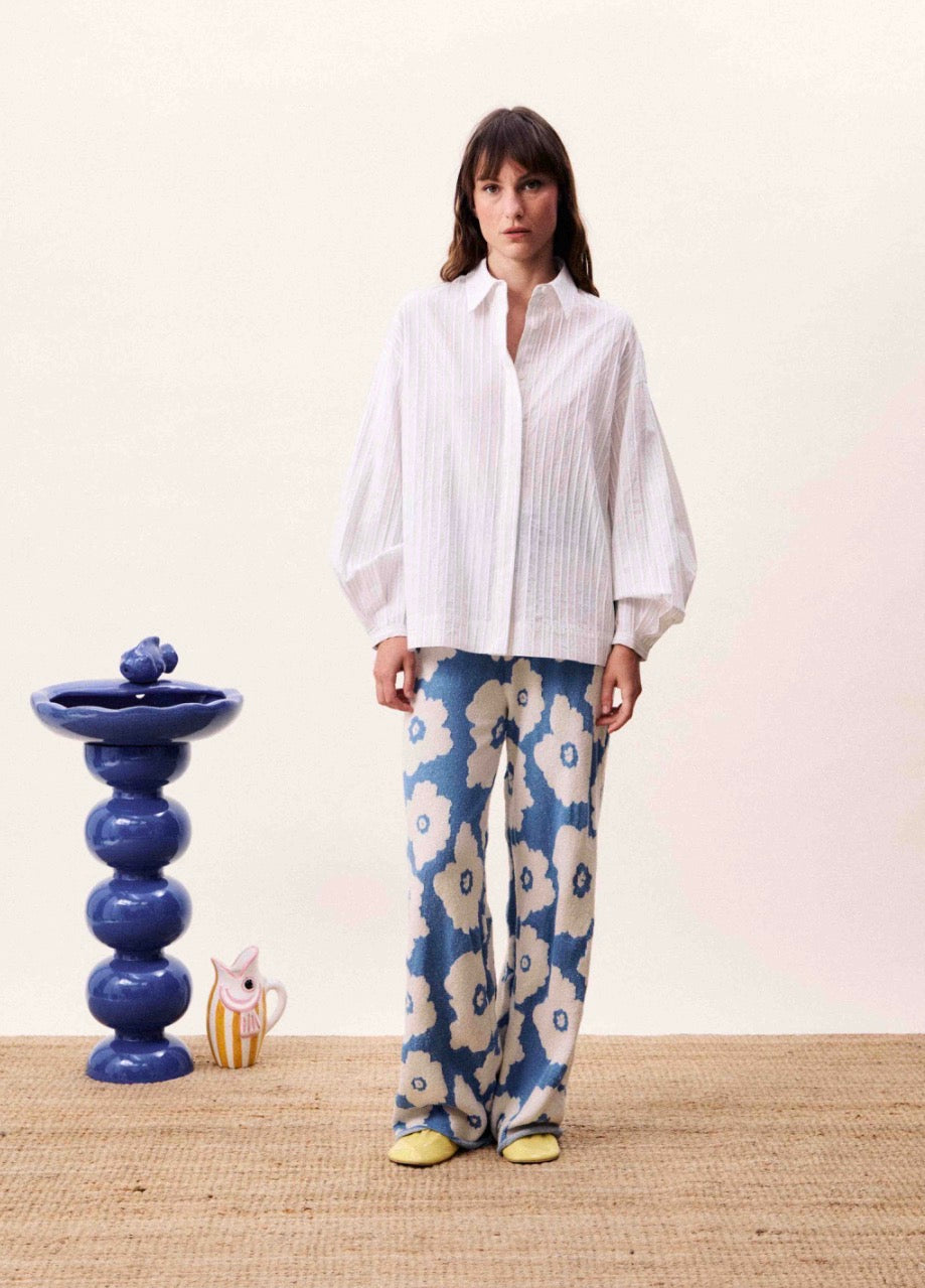 Woman wearing a white shirt and blue floral pants standing in a room with a decorative blue vase and small figurine.