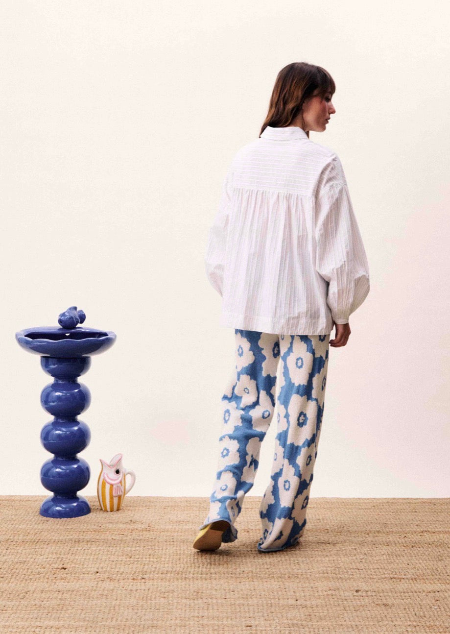 Person wearing a white blouse and blue floral pants standing in a room with a decorative blue vase and small figurine.