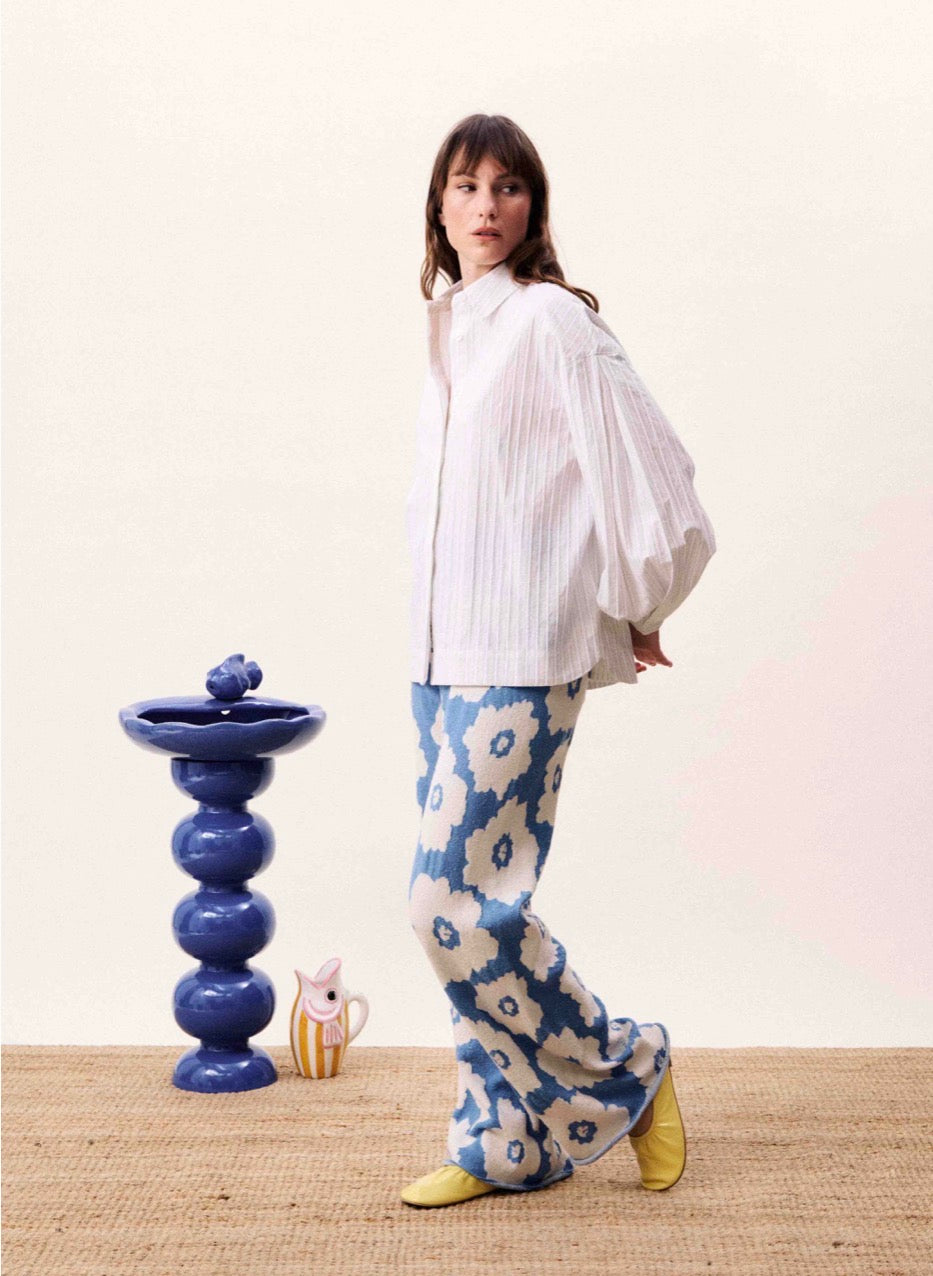 Person wearing a white blouse and blue floral pants standing next to a decorative blue vase on a beige surface.