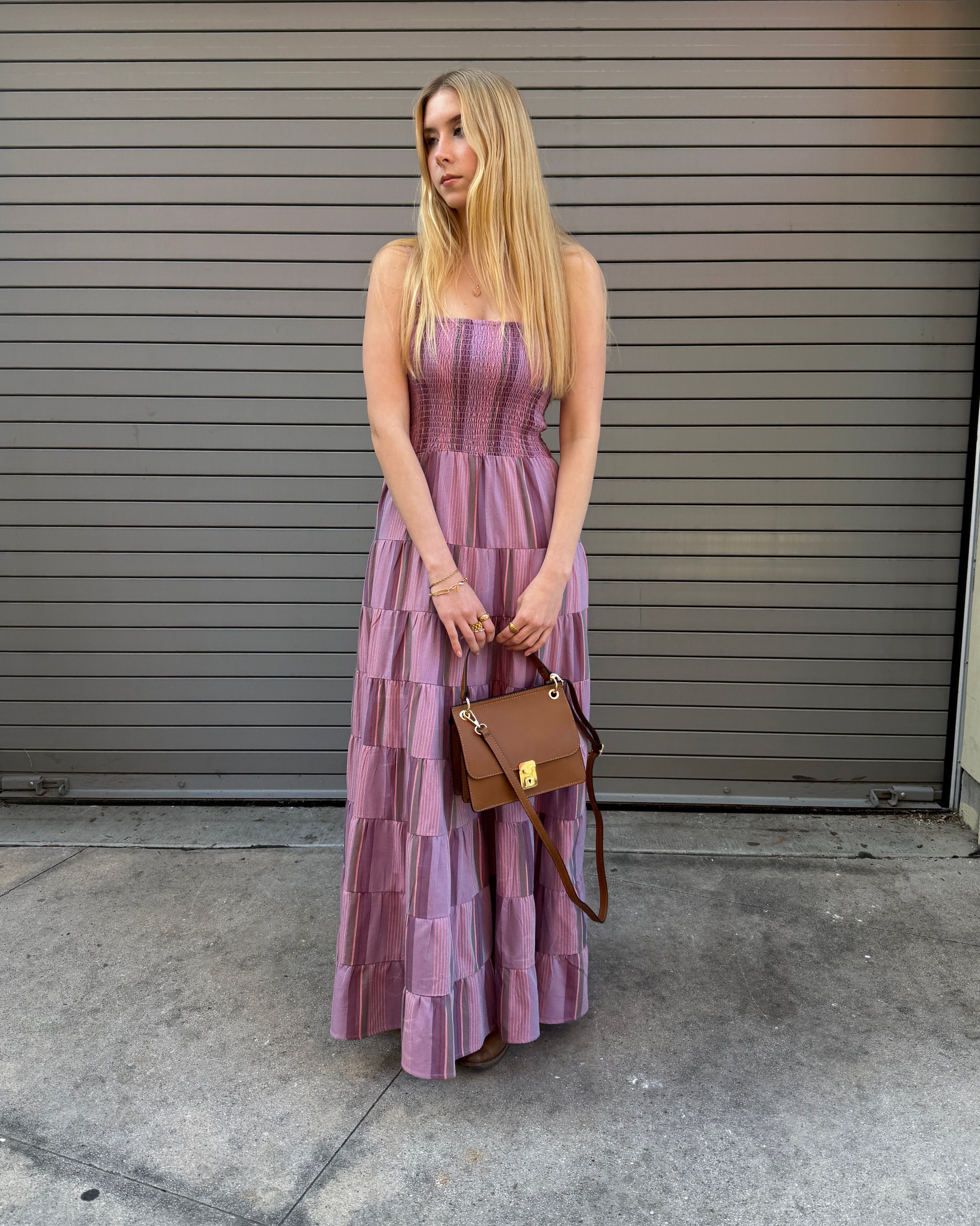 Woman in a purple dress holding a brown handbag against a gray wall.