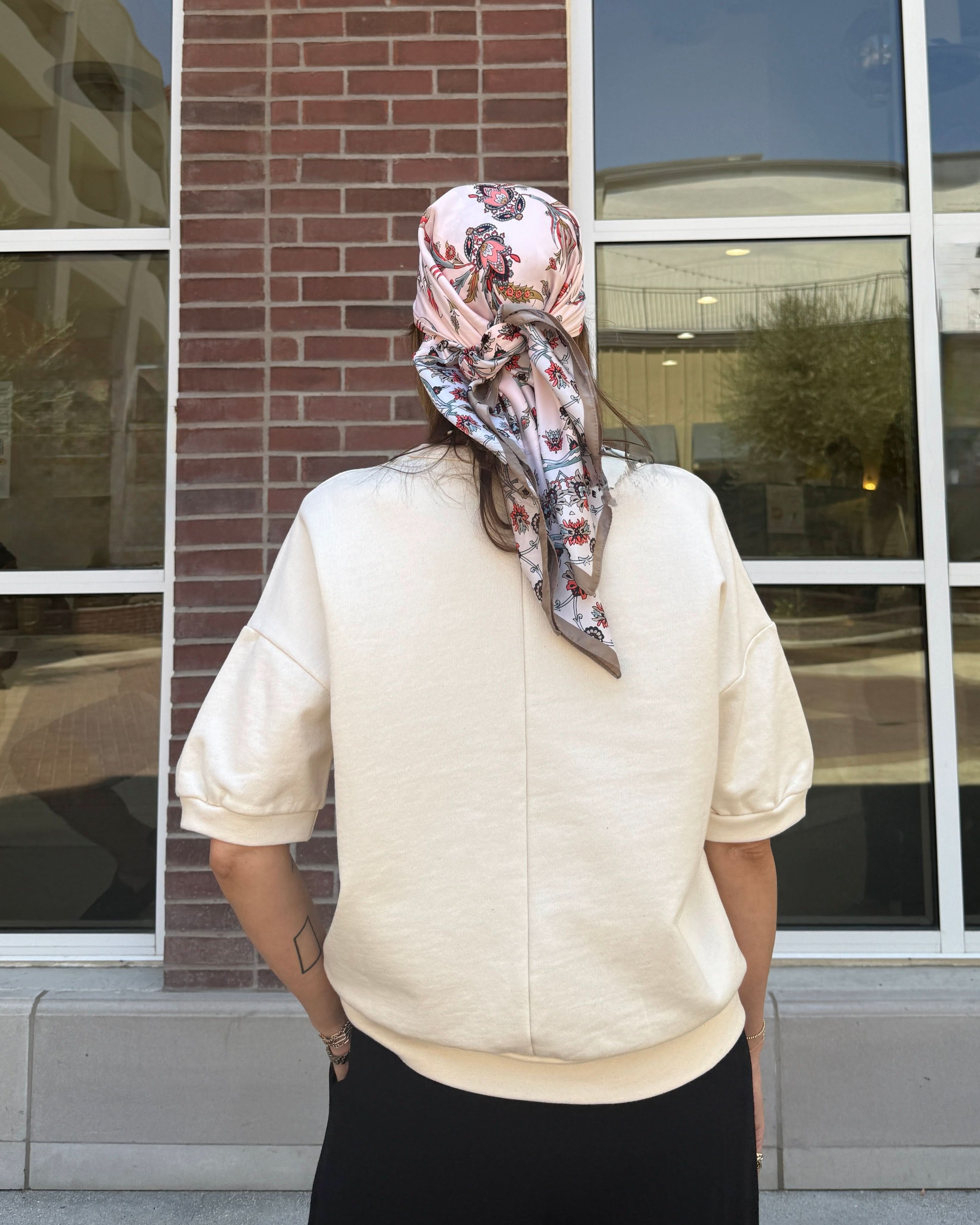 Person wearing a patterned headscarf standing in front of a building with large windows With cream short sleeve sweatshirt