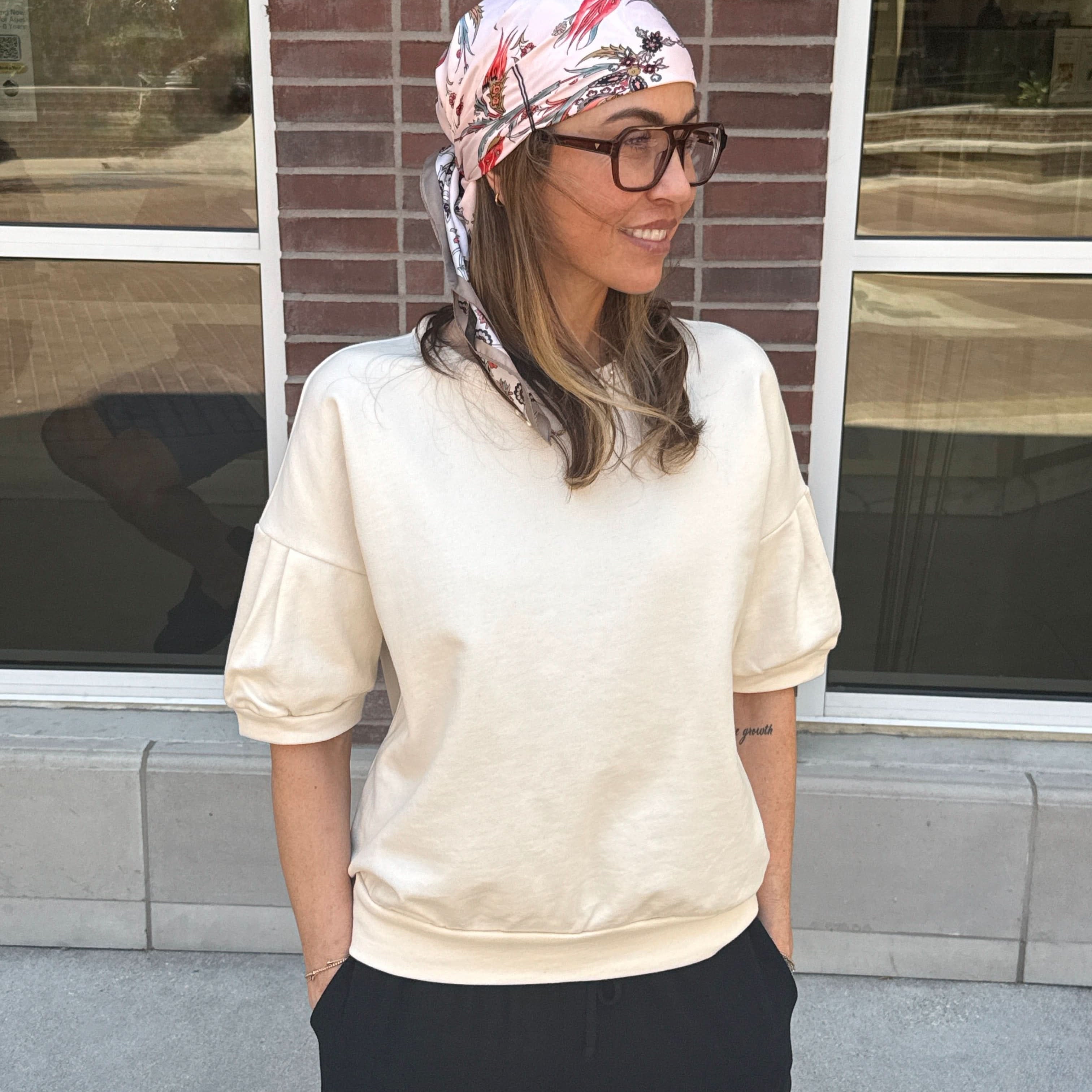 Woman wearing a headscarf and glasses standing in front of a brick wall with cream short sleeve sweatshirt