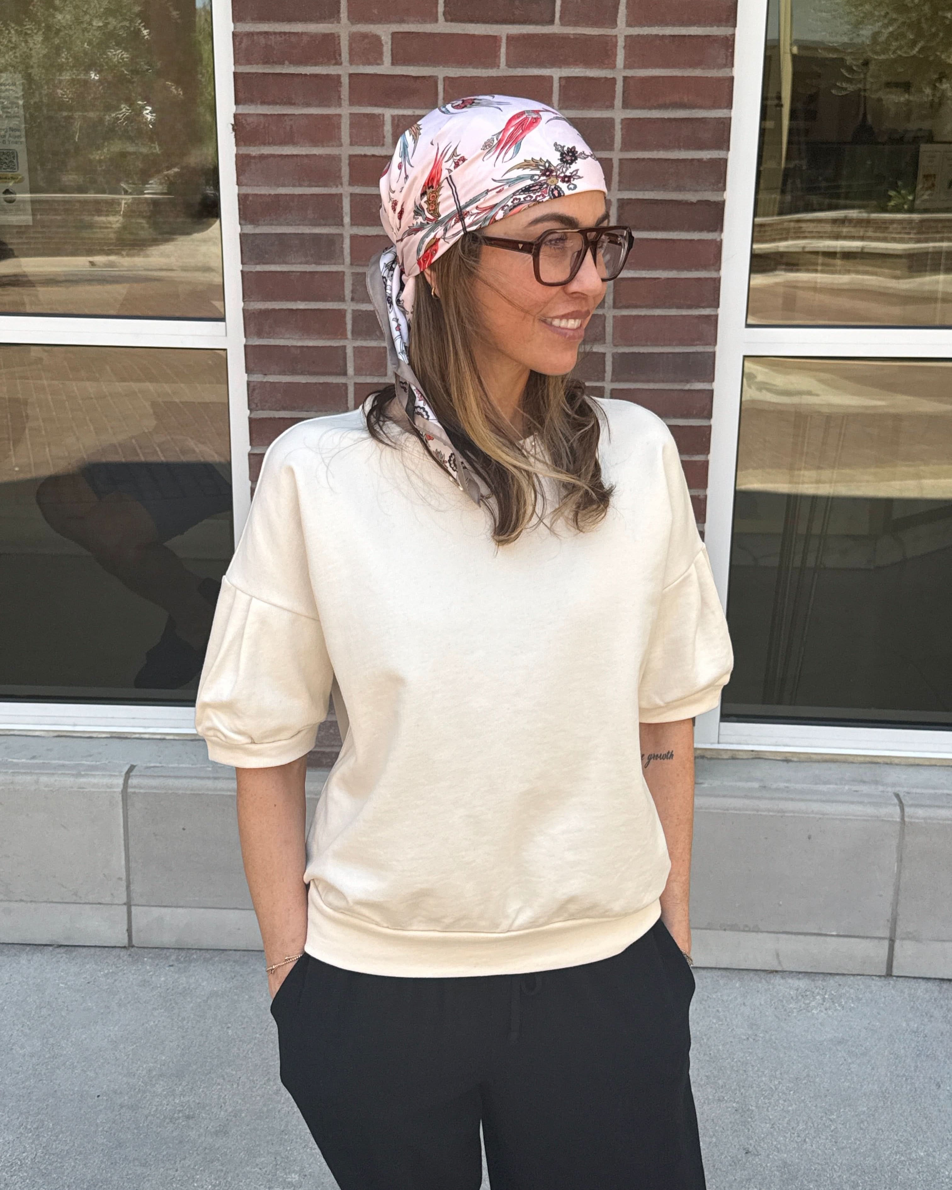 Woman wearing a headscarf and glasses standing in front of a brick wall with cream short sleeve sweatshirt