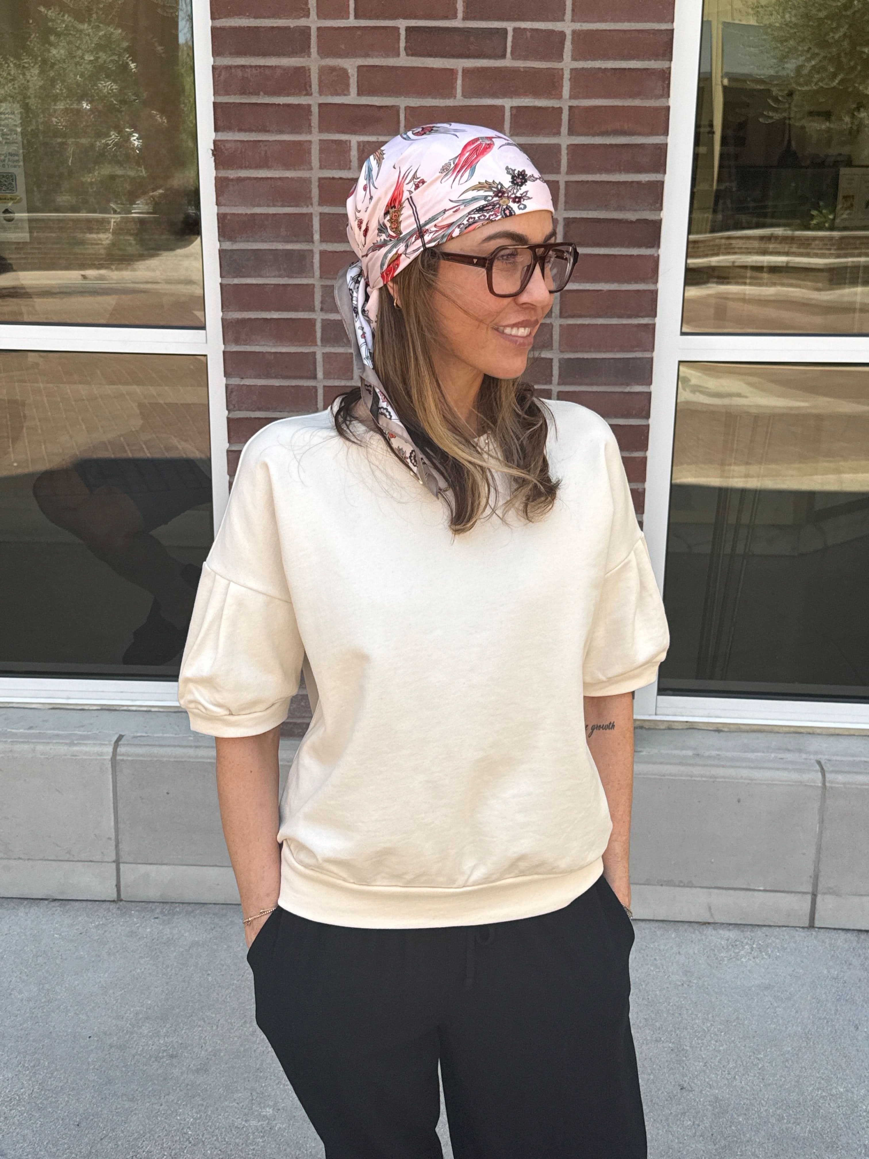 Woman wearing a headscarf and glasses standing in front of a brick wall with cream short sleeve sweatshirt
