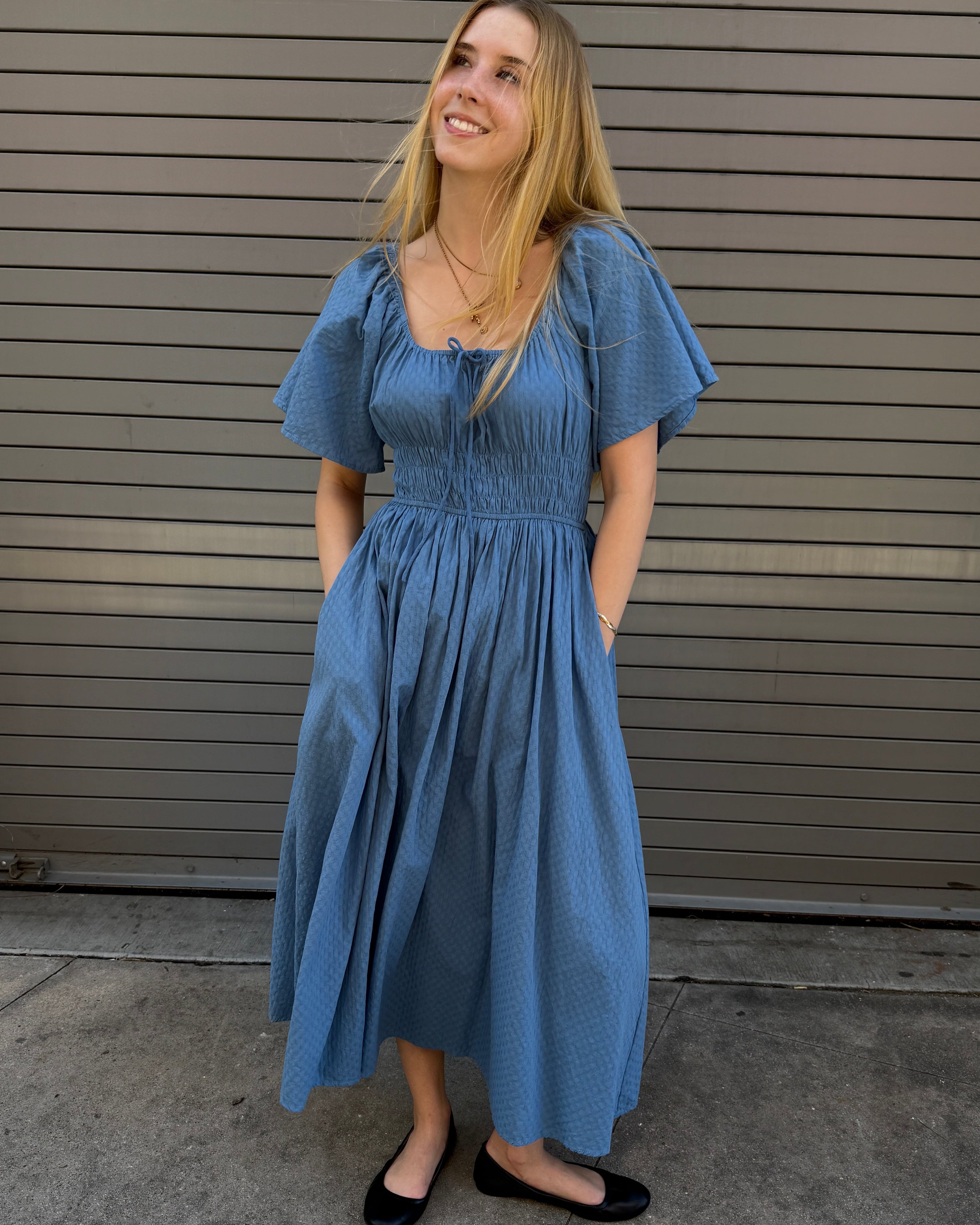 Woman wearing a blue dress standing against a gray wall.