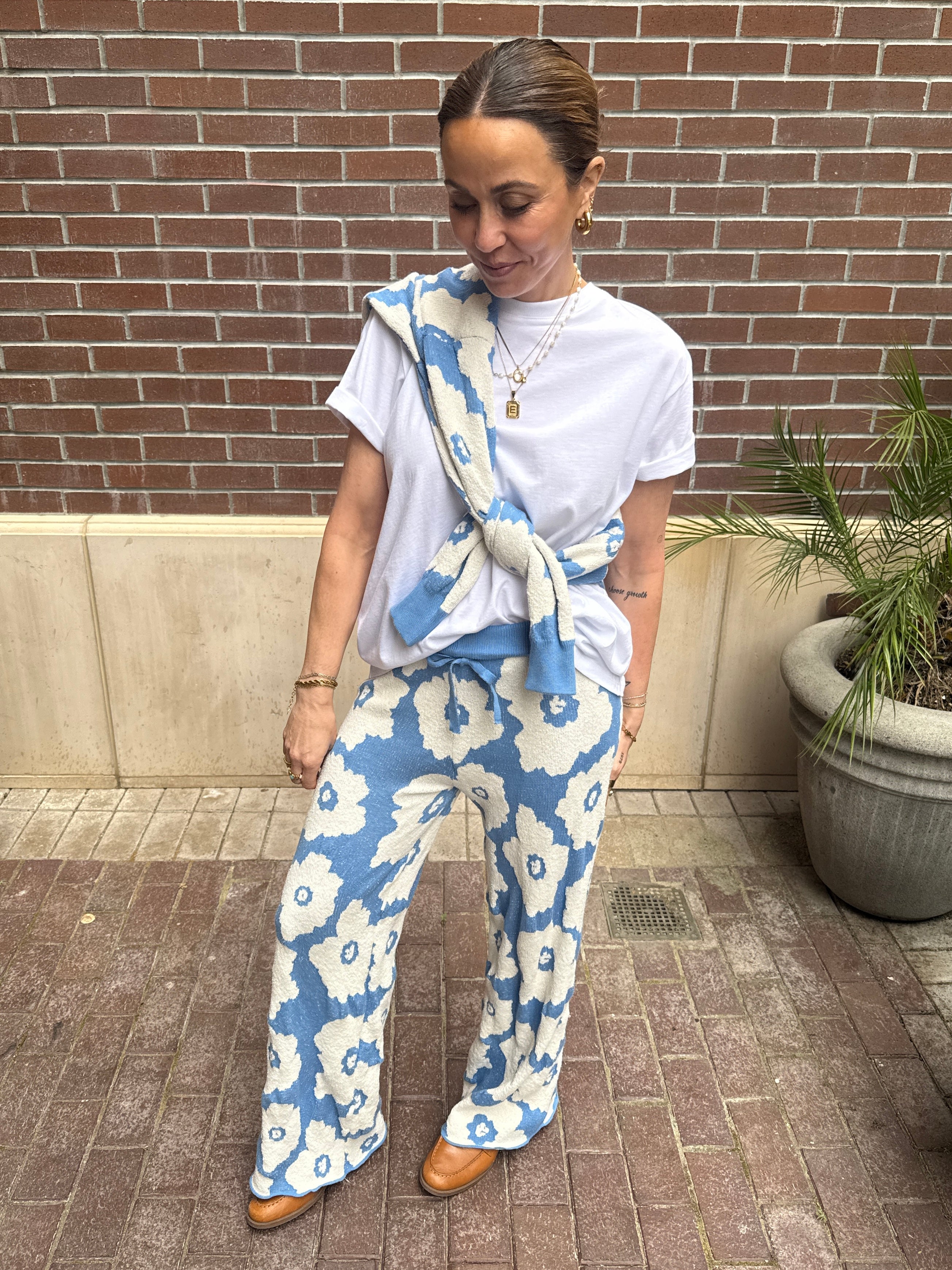 Woman wearing a white t-shirt and blue floral pants with a matching bag against a brick wall.
