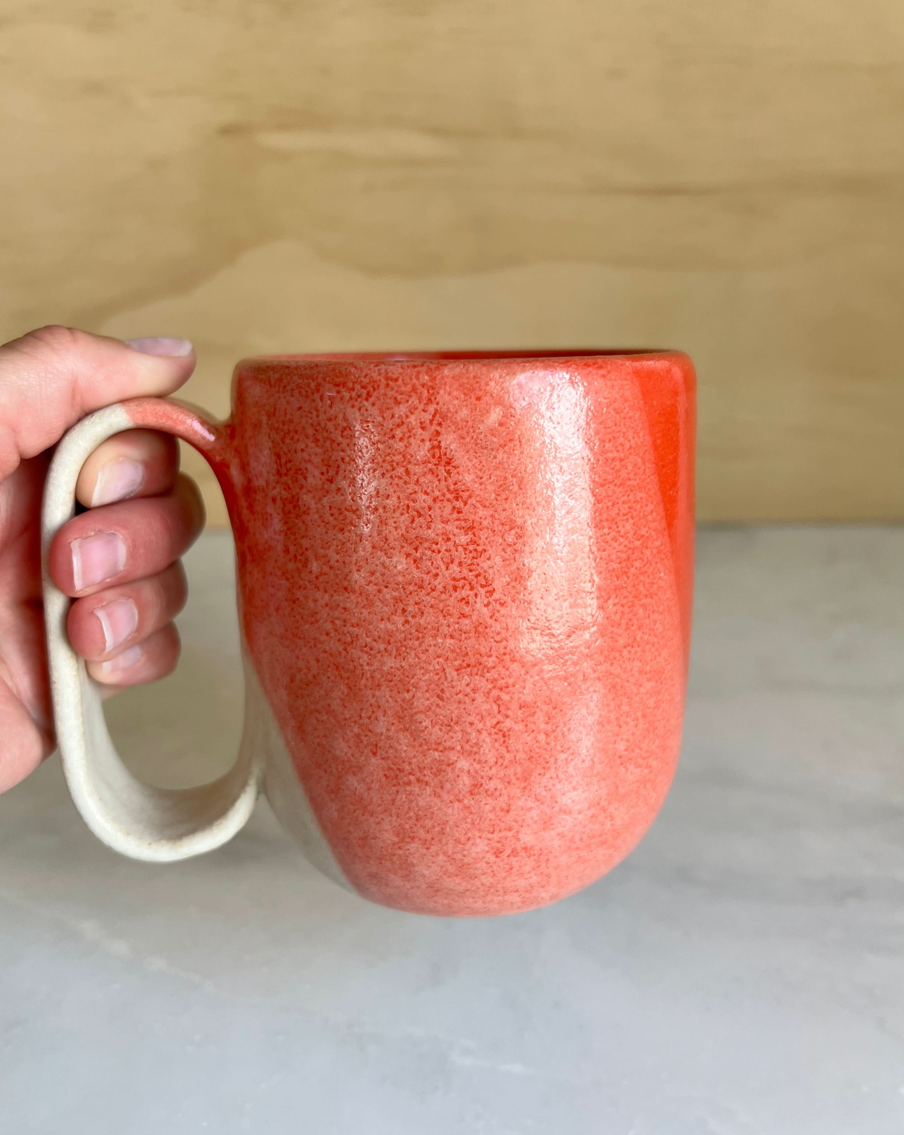 Hand holding a red ceramic mug on a light wood surface