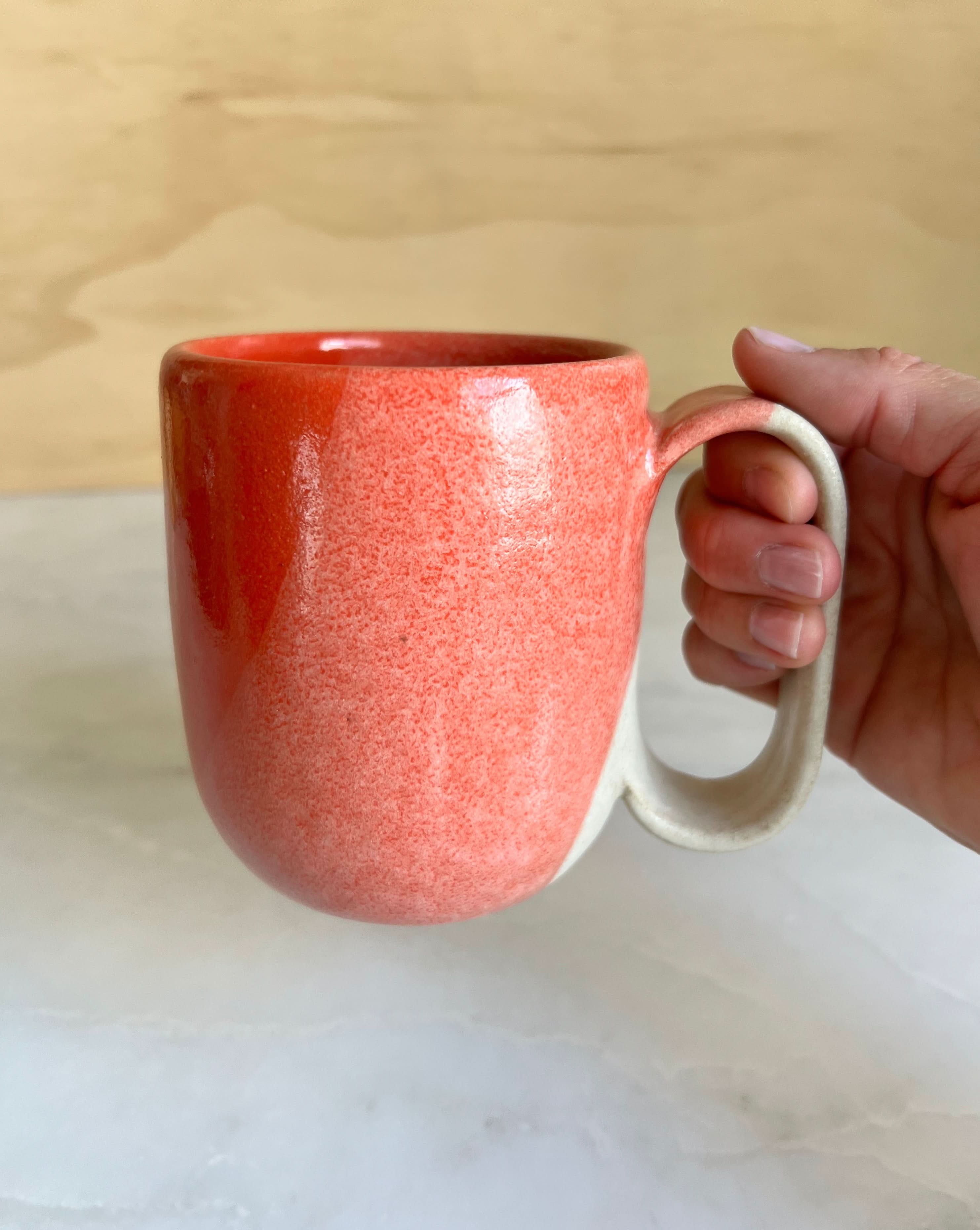 Hand holding a red ceramic mug on a light-colored surface with a wooden background