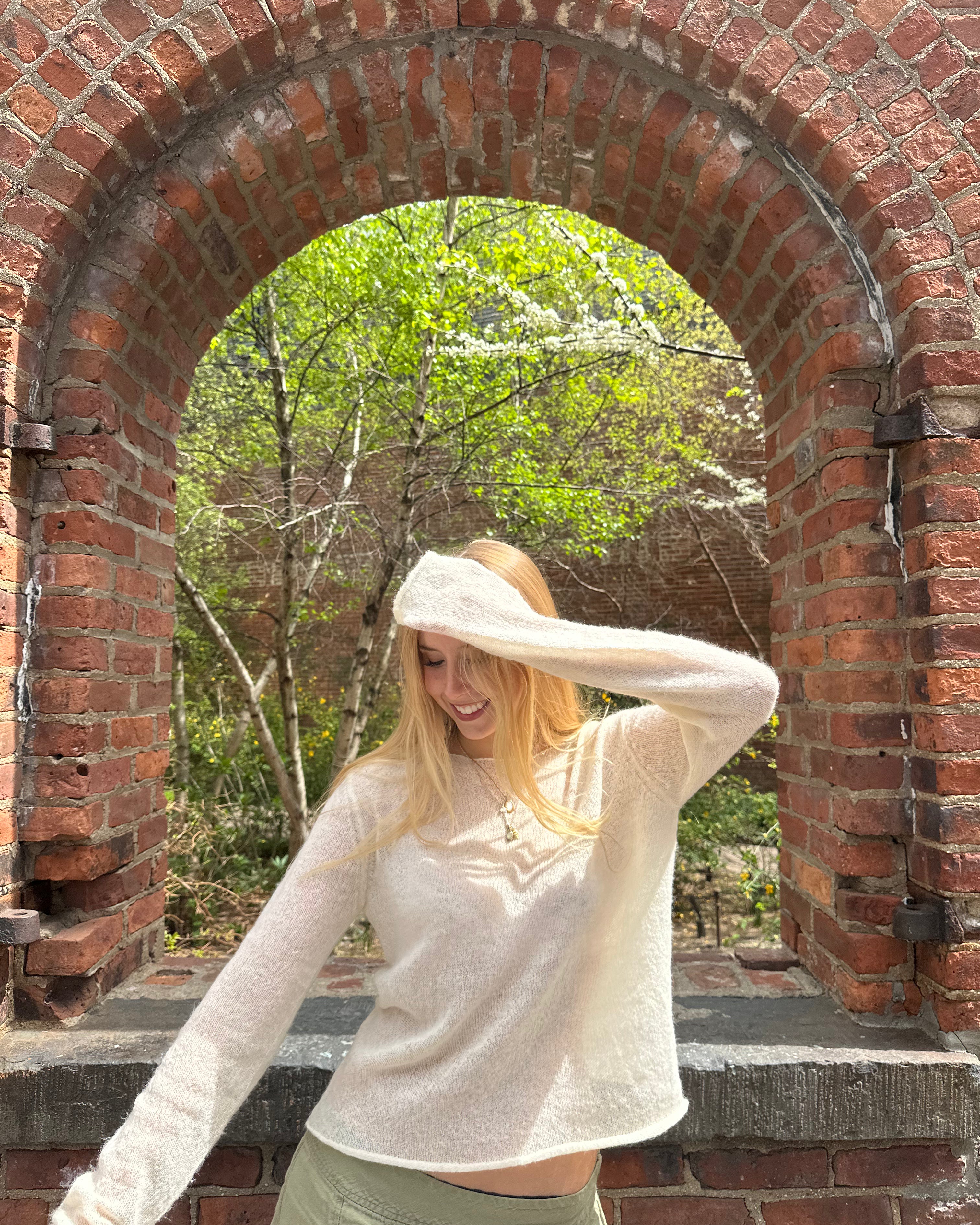 Woman standing under a brick archway with trees in the background wearing cream sweater