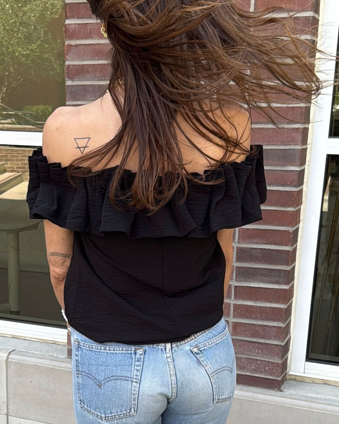 Woman wearing a black off the shoulder ruffle top with blue jeans with pockets behind a brick wall.