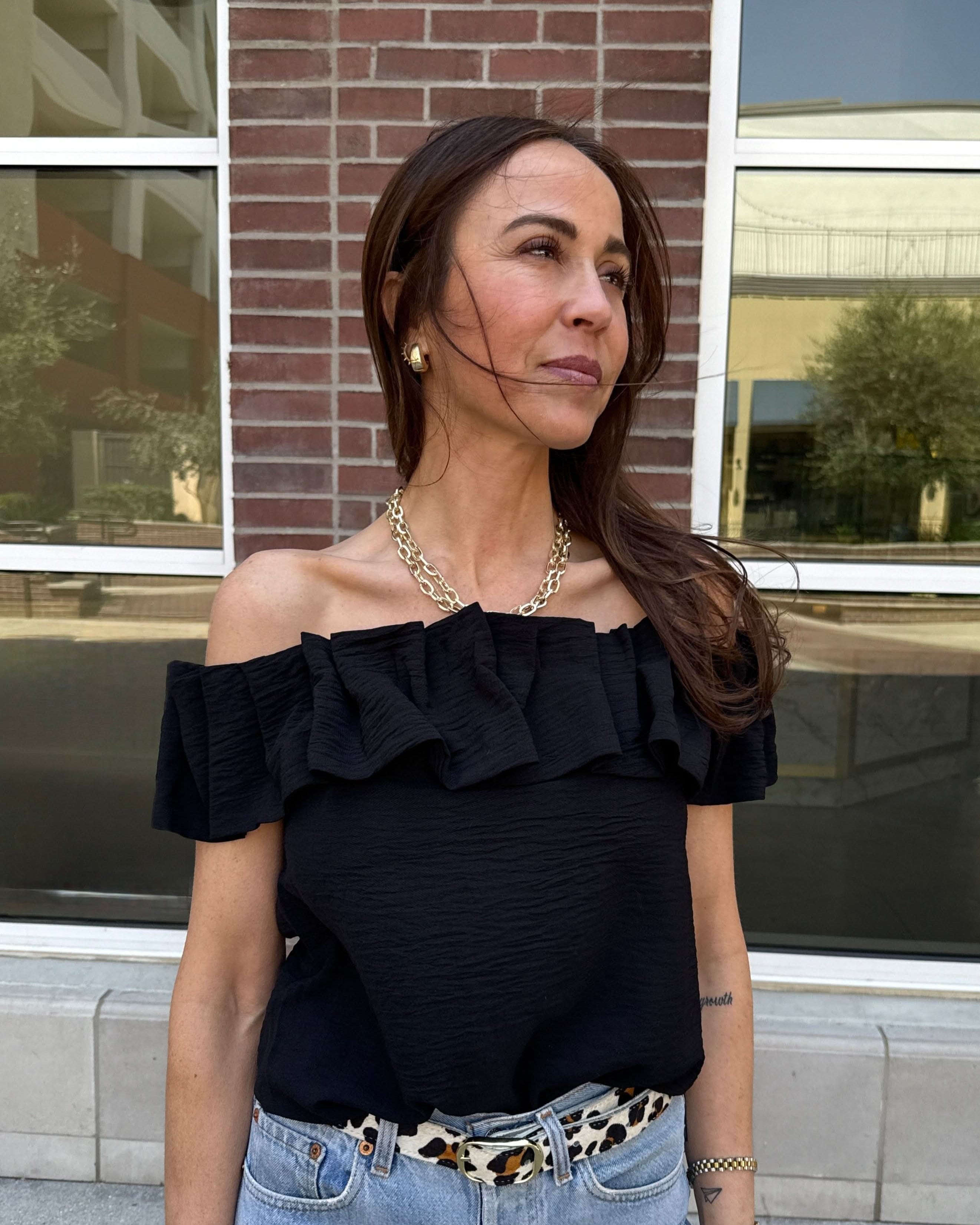 Woman wearing a black off the shoulder ruffle top behind a brick wall.