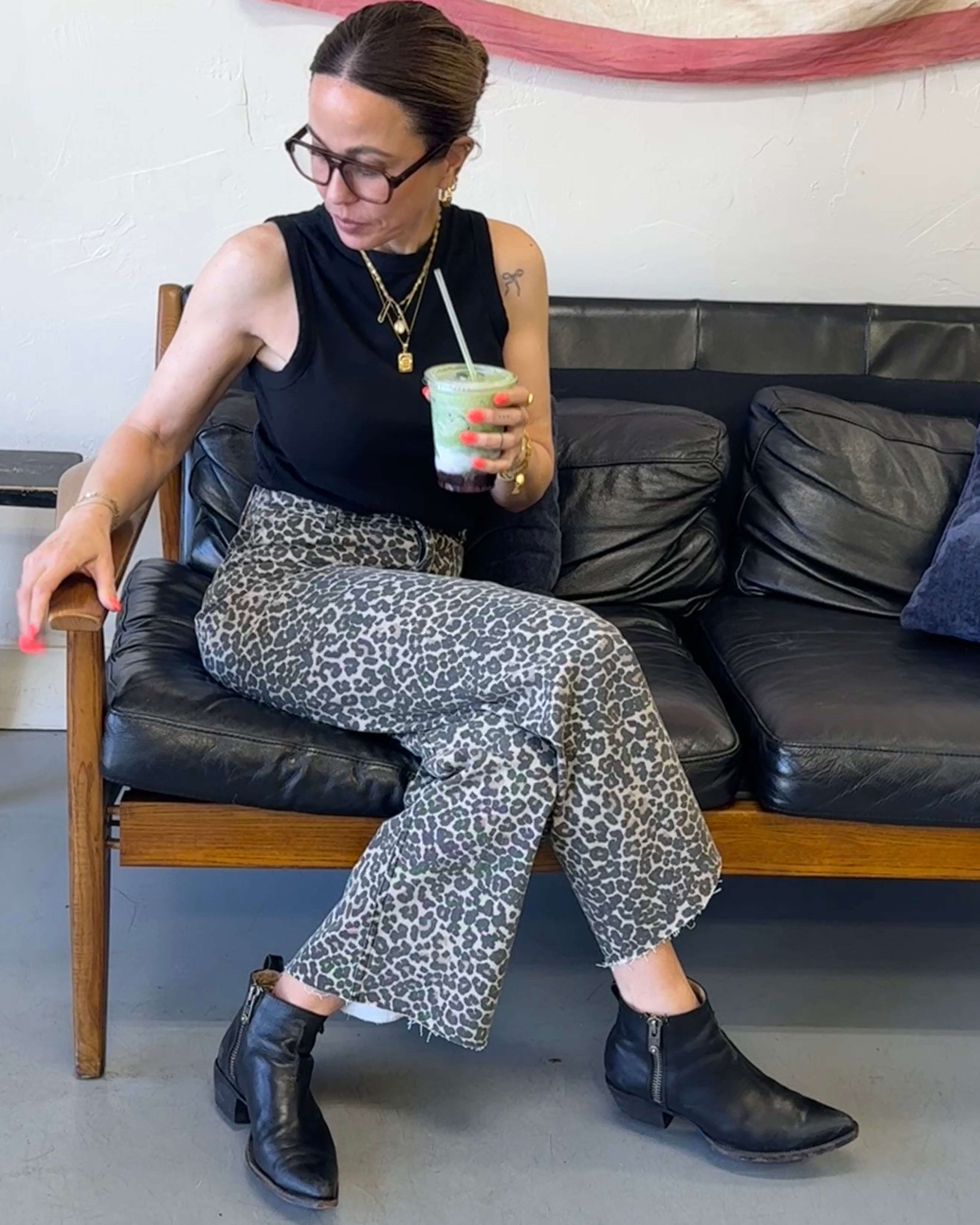 Woman sitting on a black leather couch holding a drink, wearing a black sleeveless top and patterned pants.
