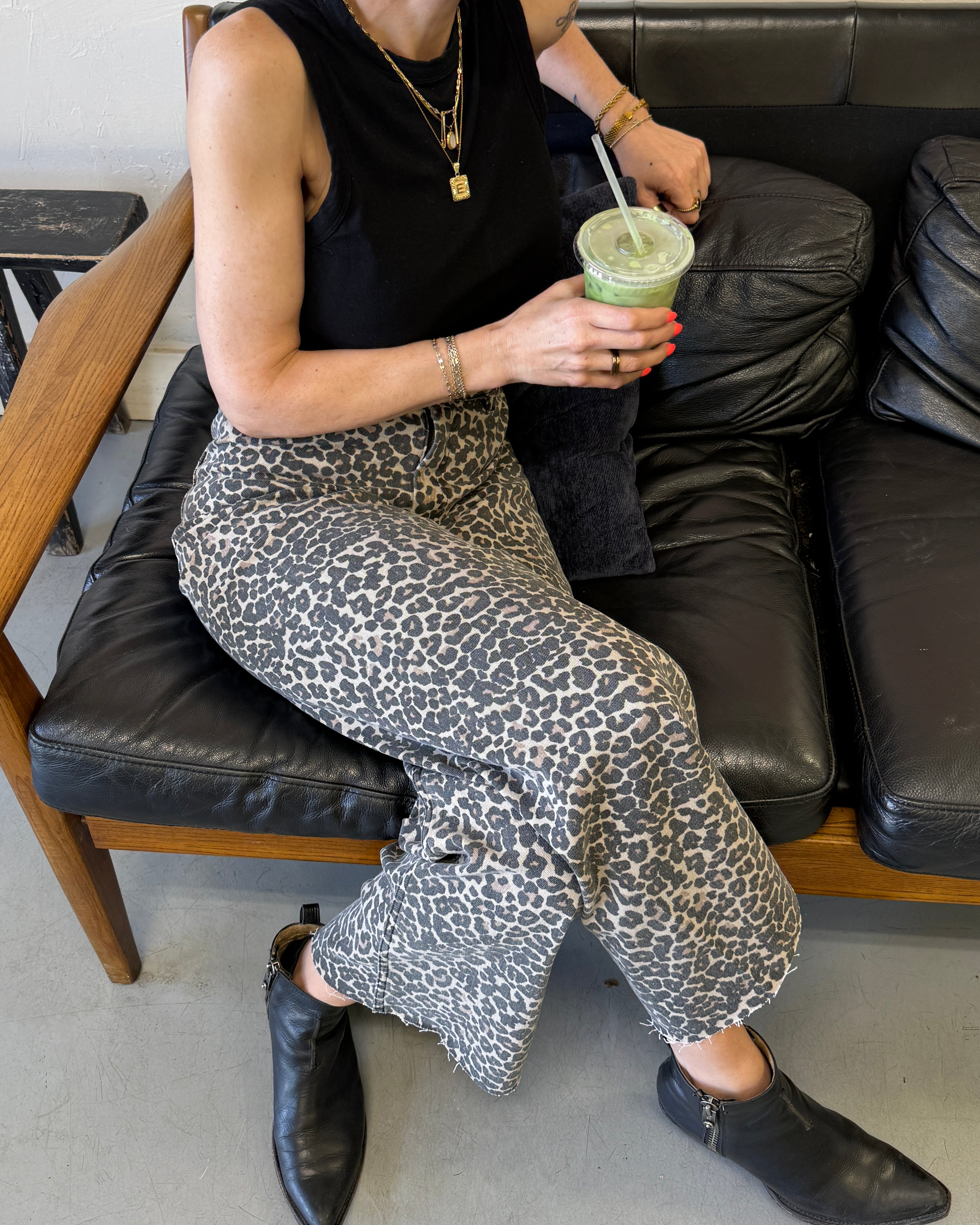 Person sitting on a couch holding a green drink, wearing a black sleeveless top and leopard print pants.