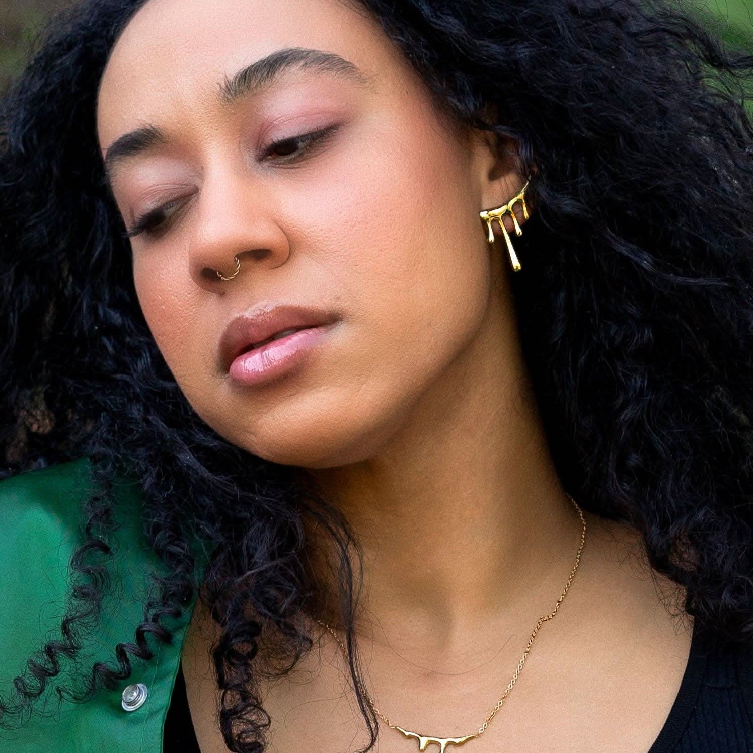 Woman with curly hair wearing gold earrings and a necklace, with a blurred natural background