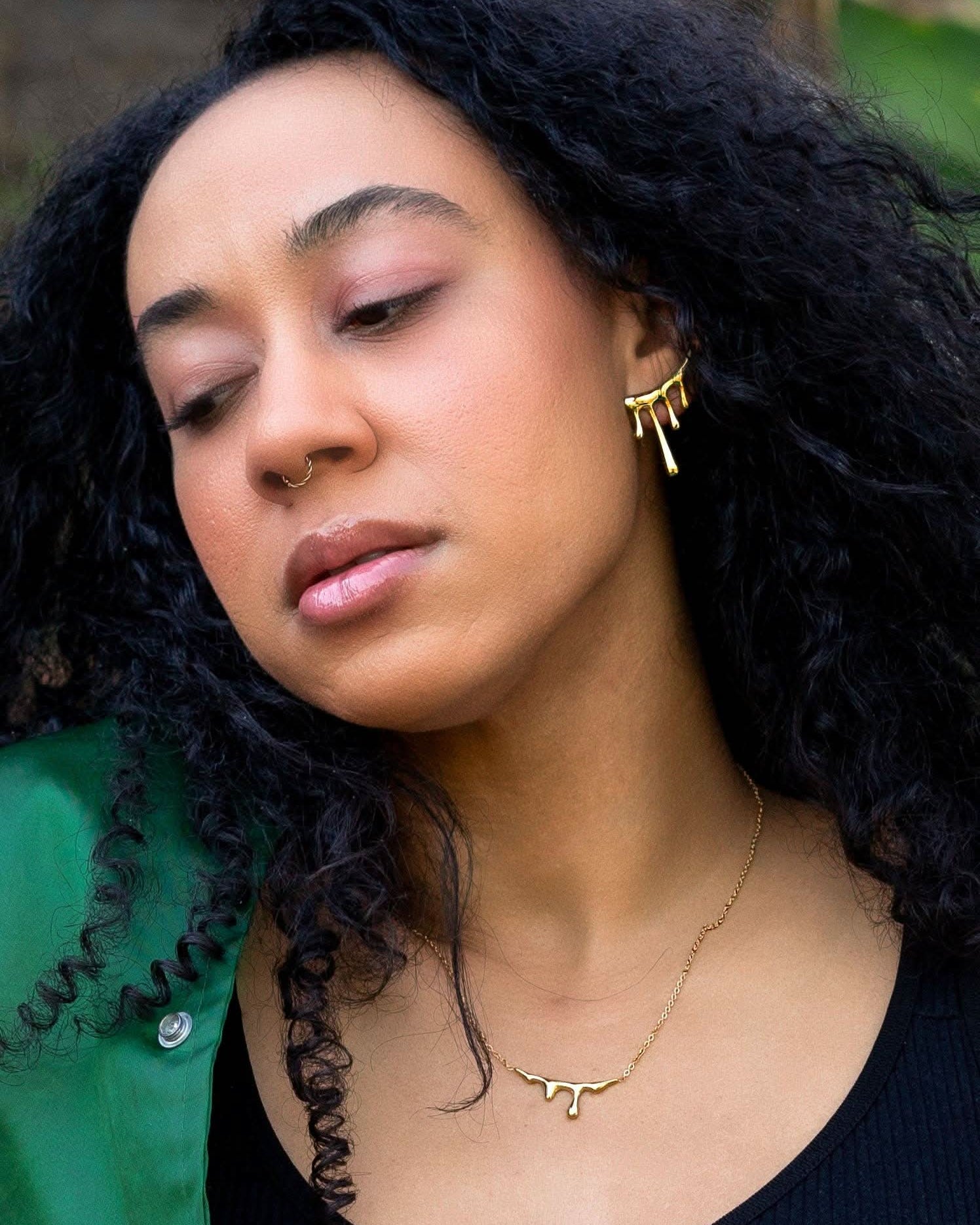 Woman with curly hair wearing gold earrings and a necklace, with a blurred natural background