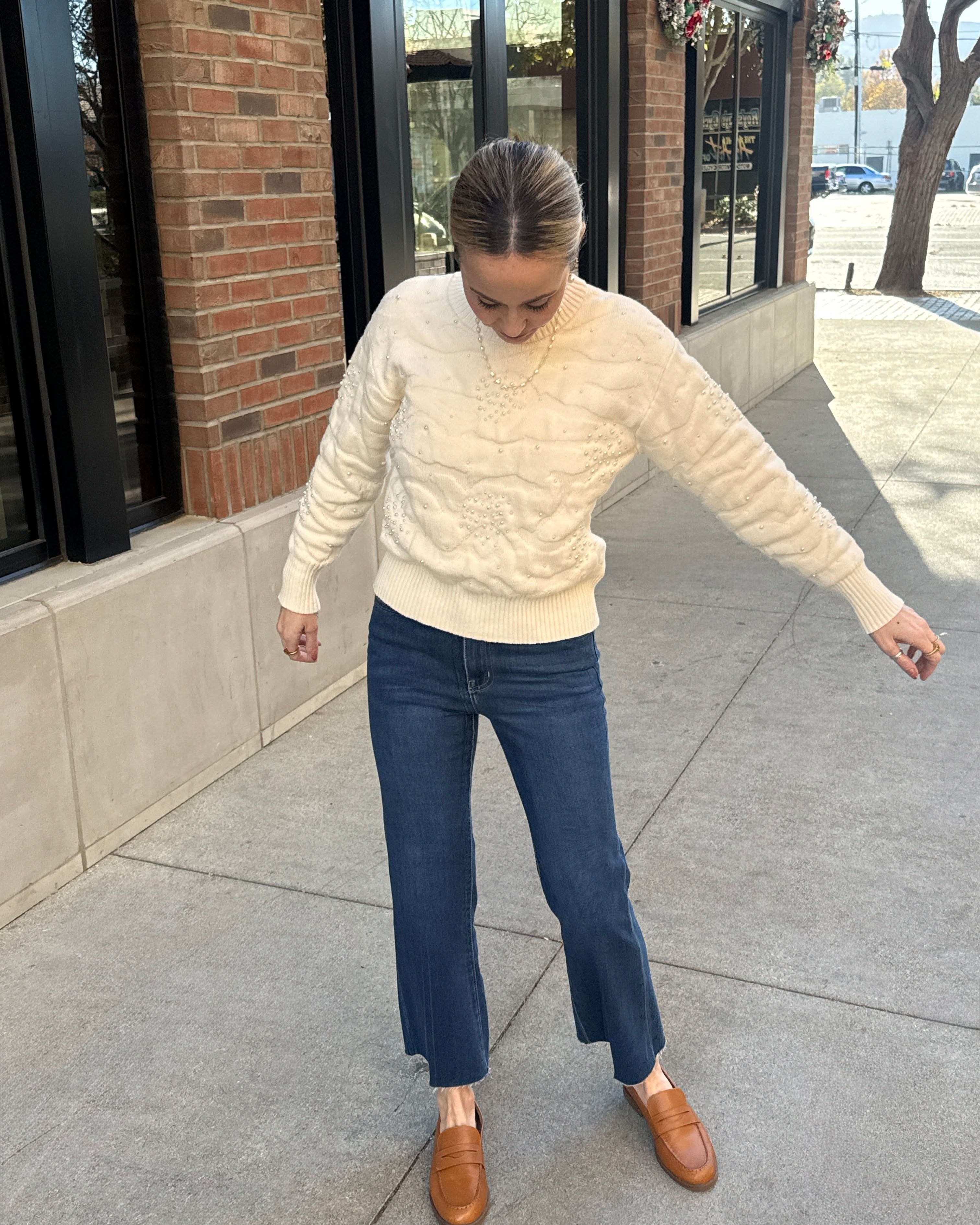 Person wearing a cream sweater, blue jeans, and brown shoes on a sidewalk.