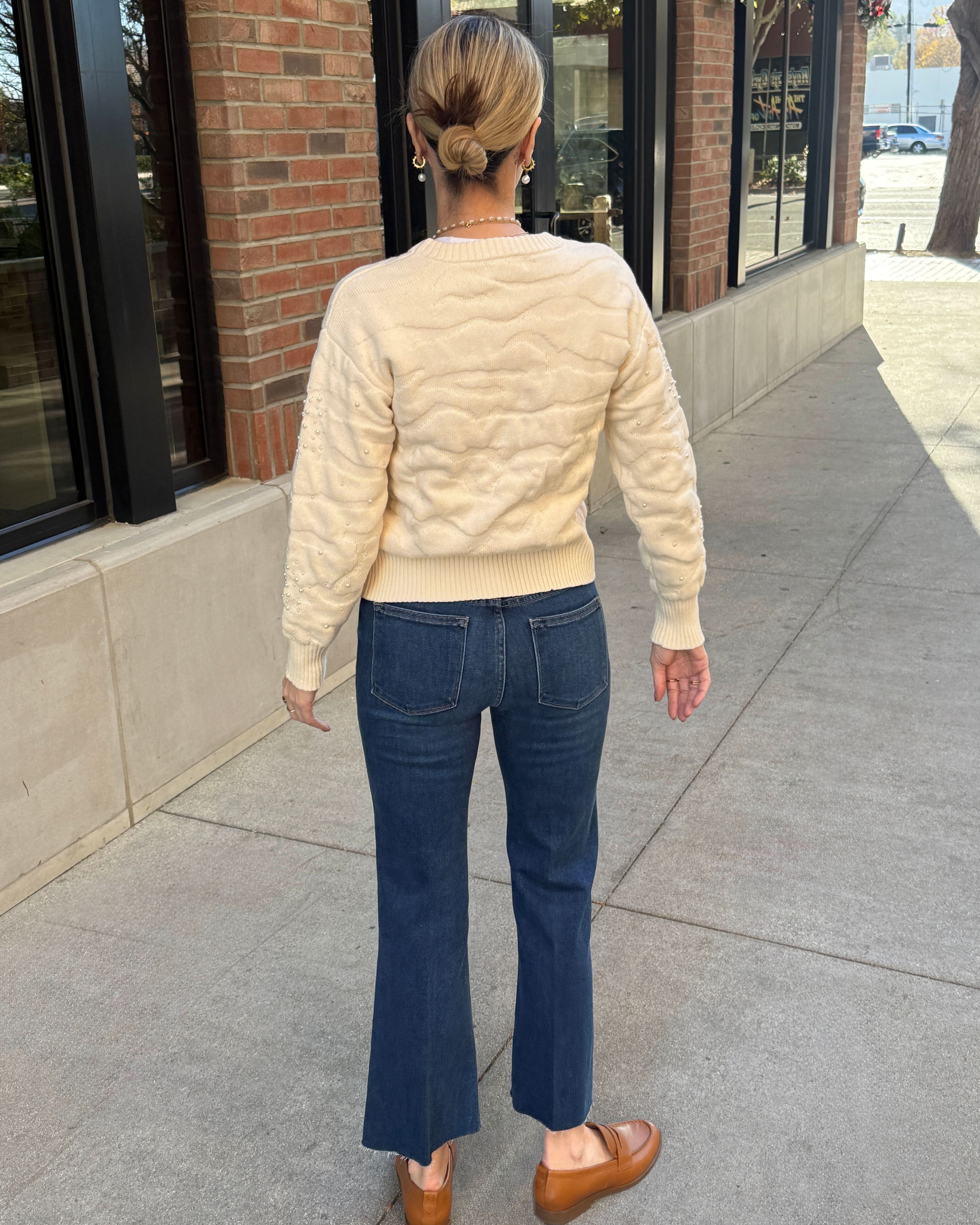 Person wearing a cream quilted sweater and blue jeans standing on a sidewalk.