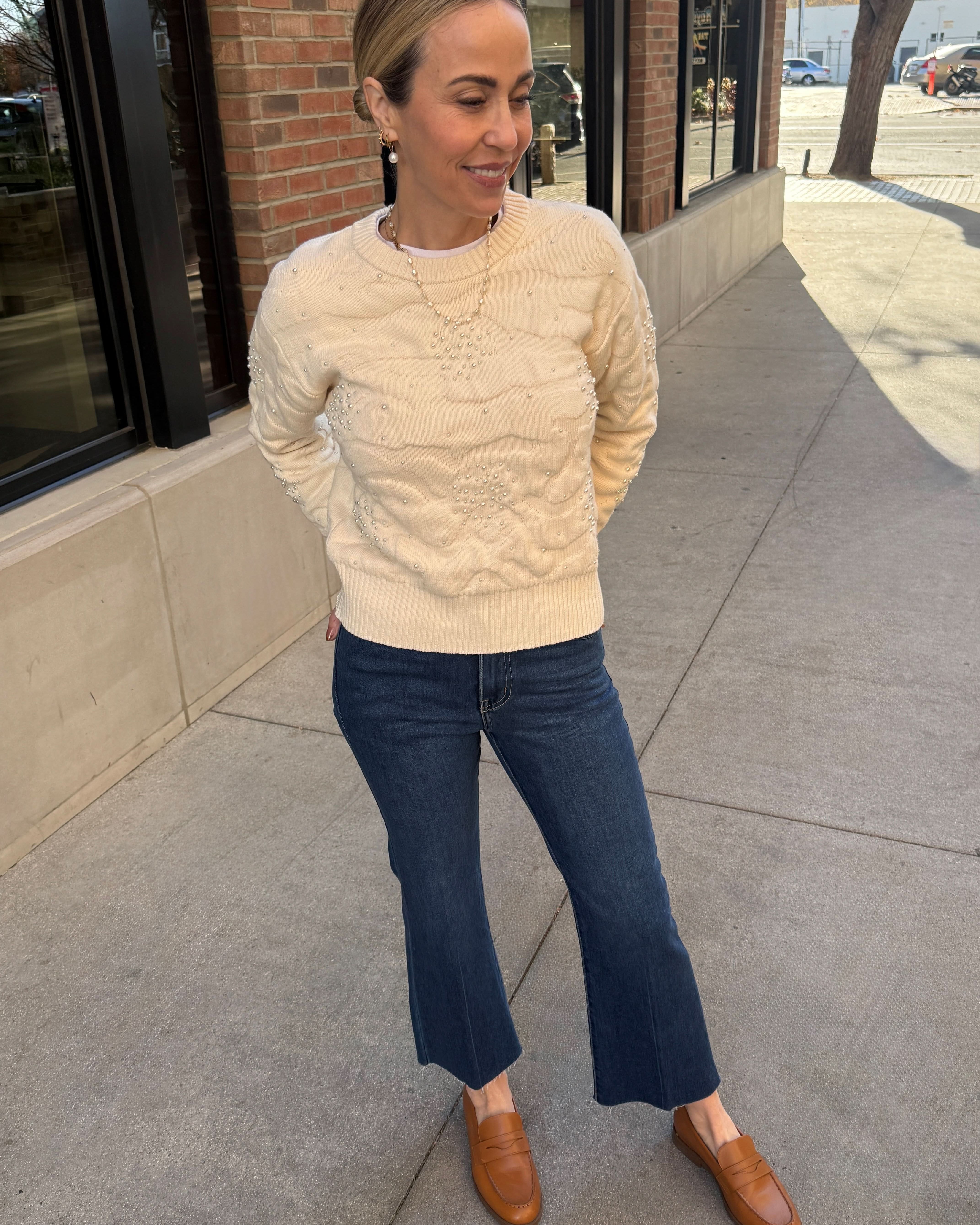 Woman wearing a beige sweater and blue jeans standing on a sidewalk.