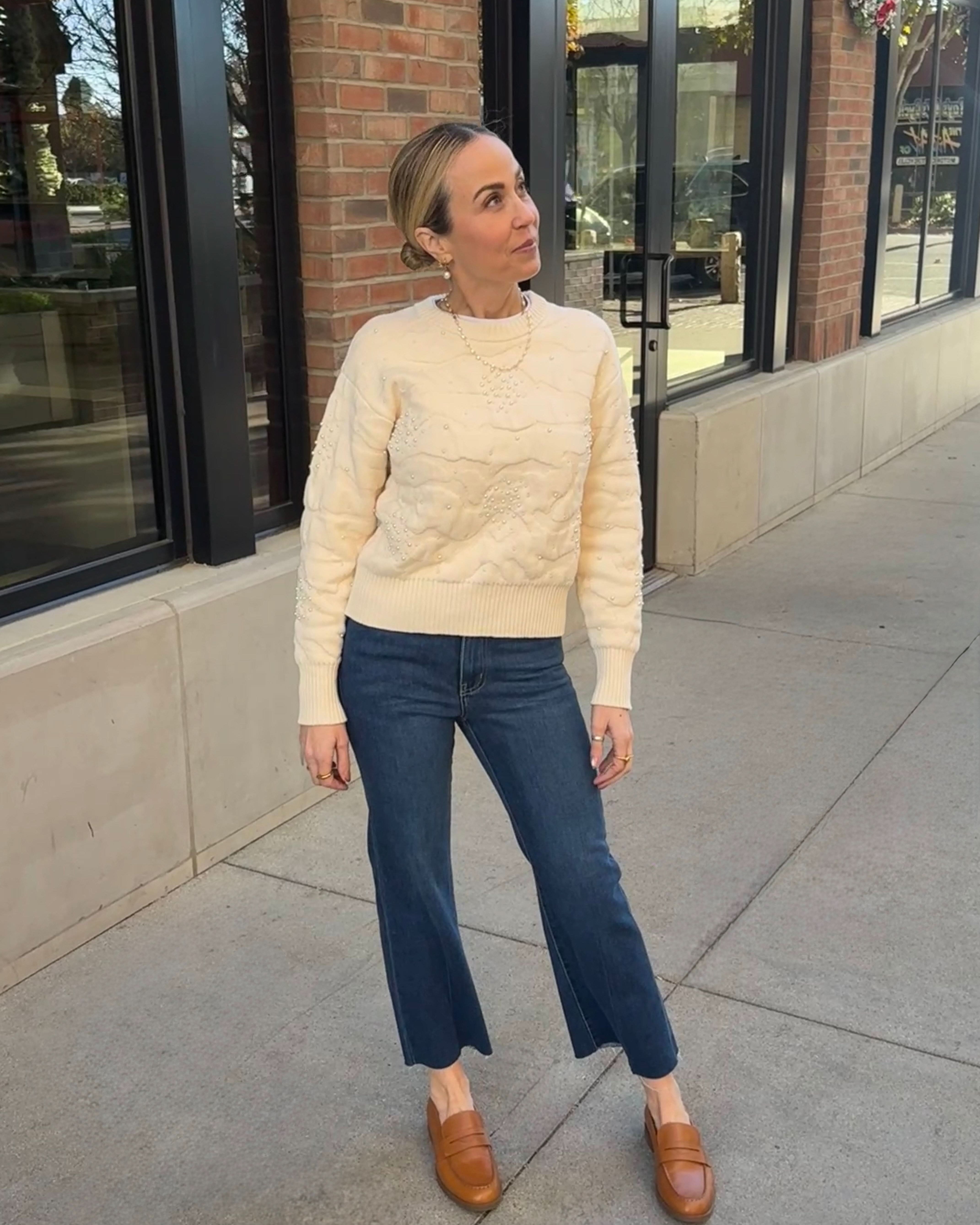 Woman wearing a cream sweater and blue jeans standing on a sidewalk.