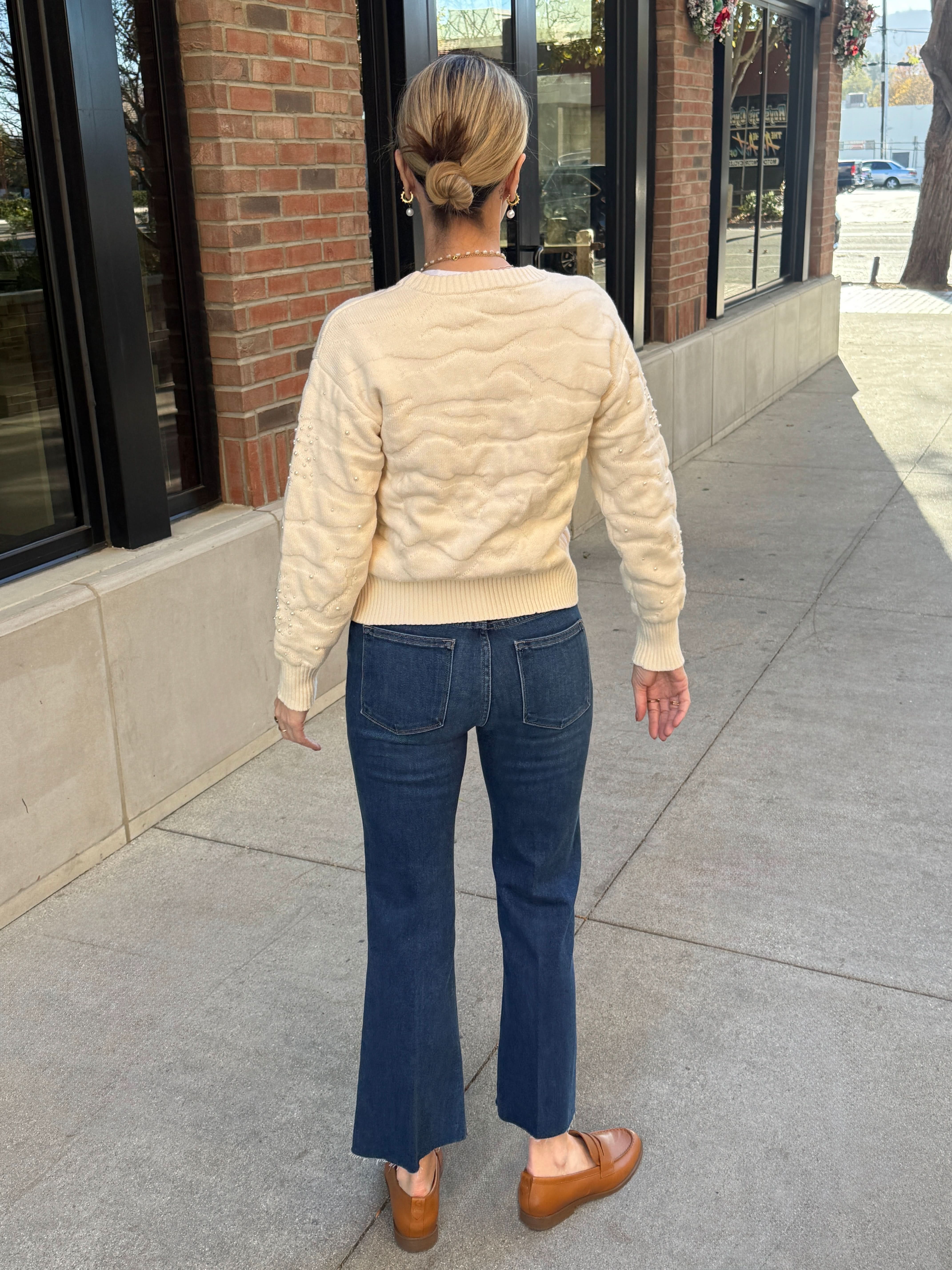 Person wearing a cream quilted sweater and blue jeans standing on a sidewalk.
