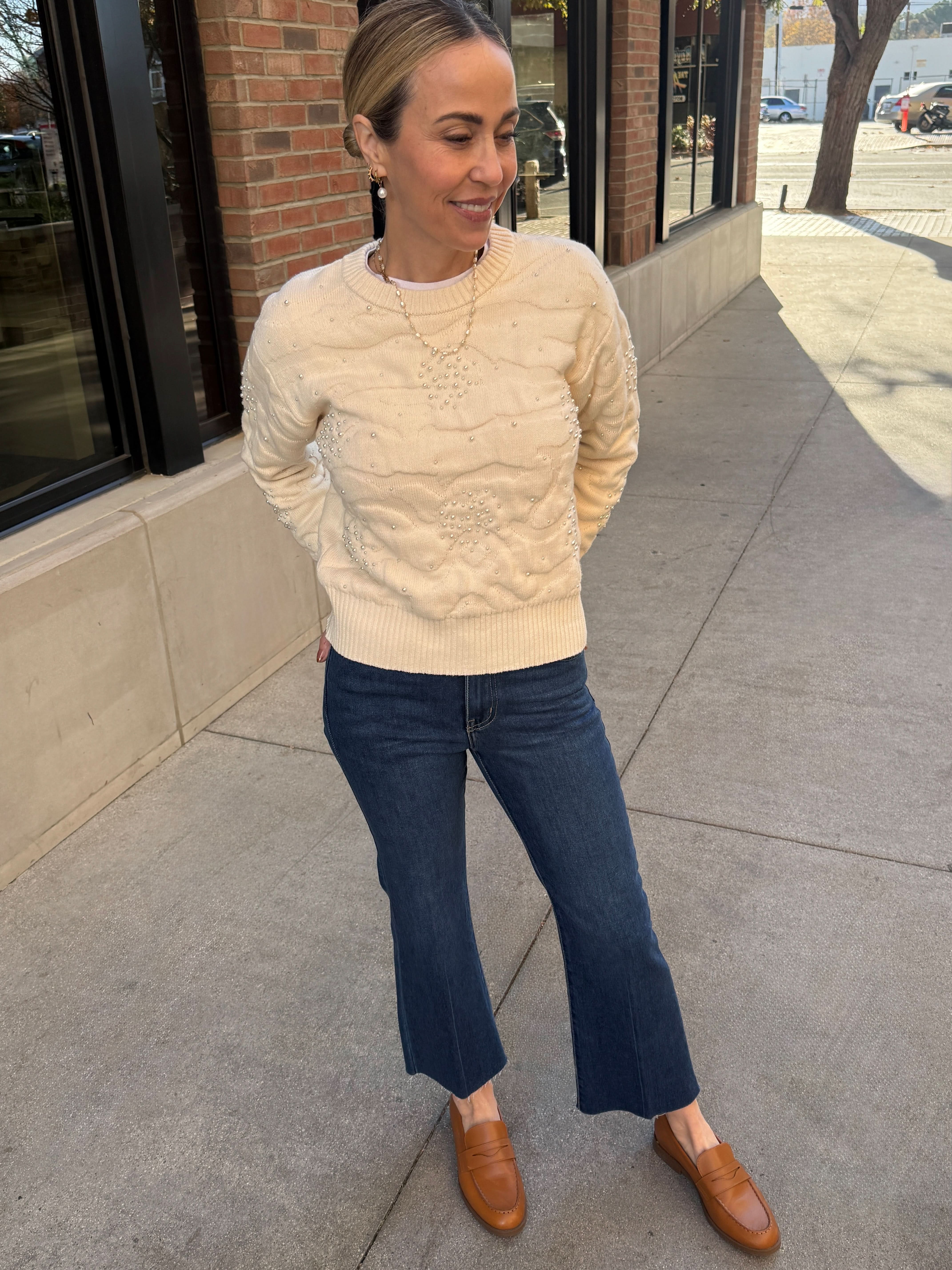 Woman wearing a beige sweater and blue jeans standing on a sidewalk.