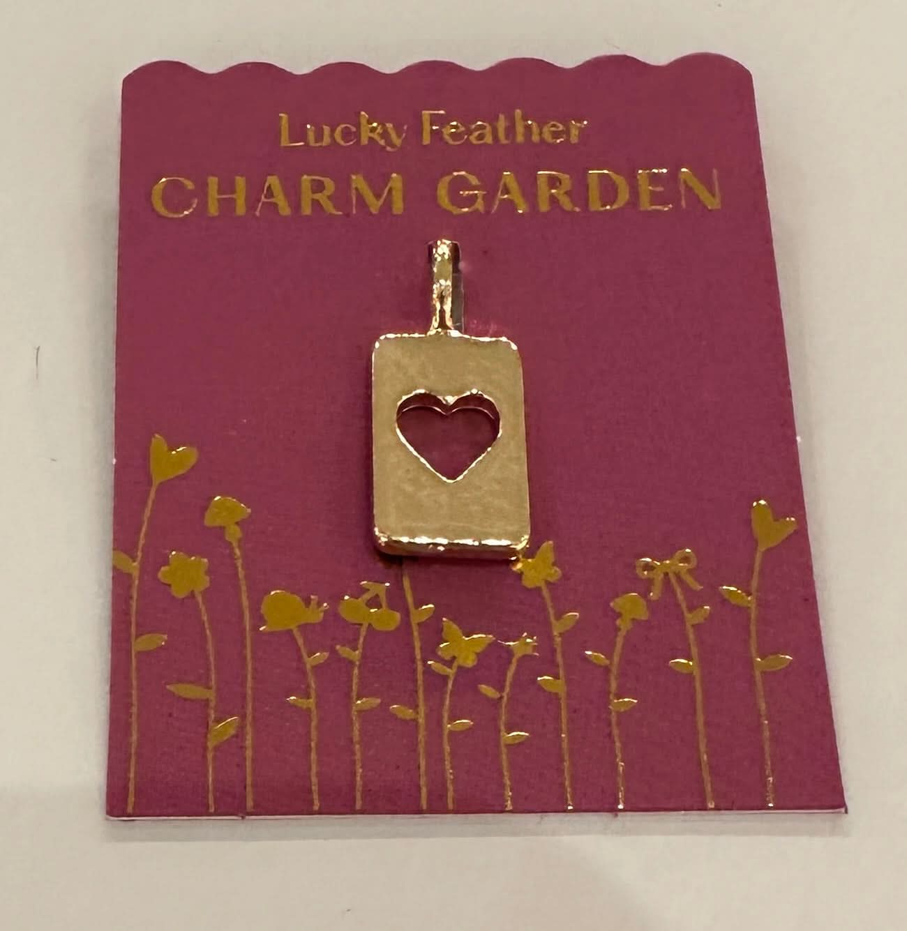 Gold charm with heart cutout on a 'Lucky Feather Charm Garden' card with floral design