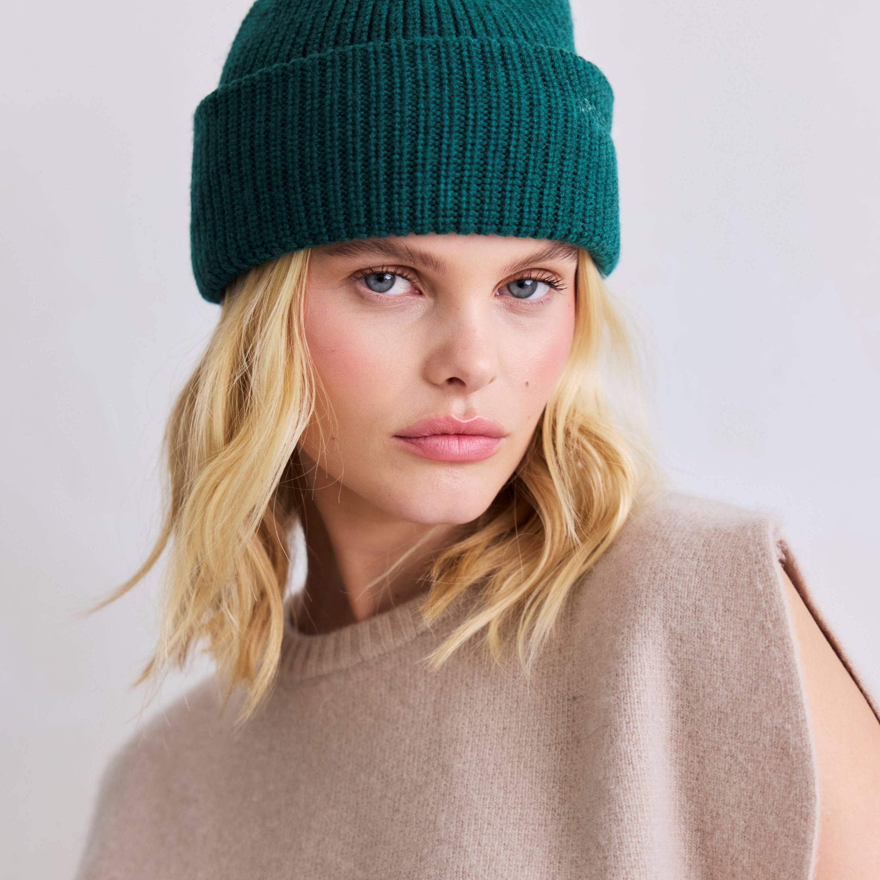 Woman wearing a teal beanie and beige sleeveless top against a white background