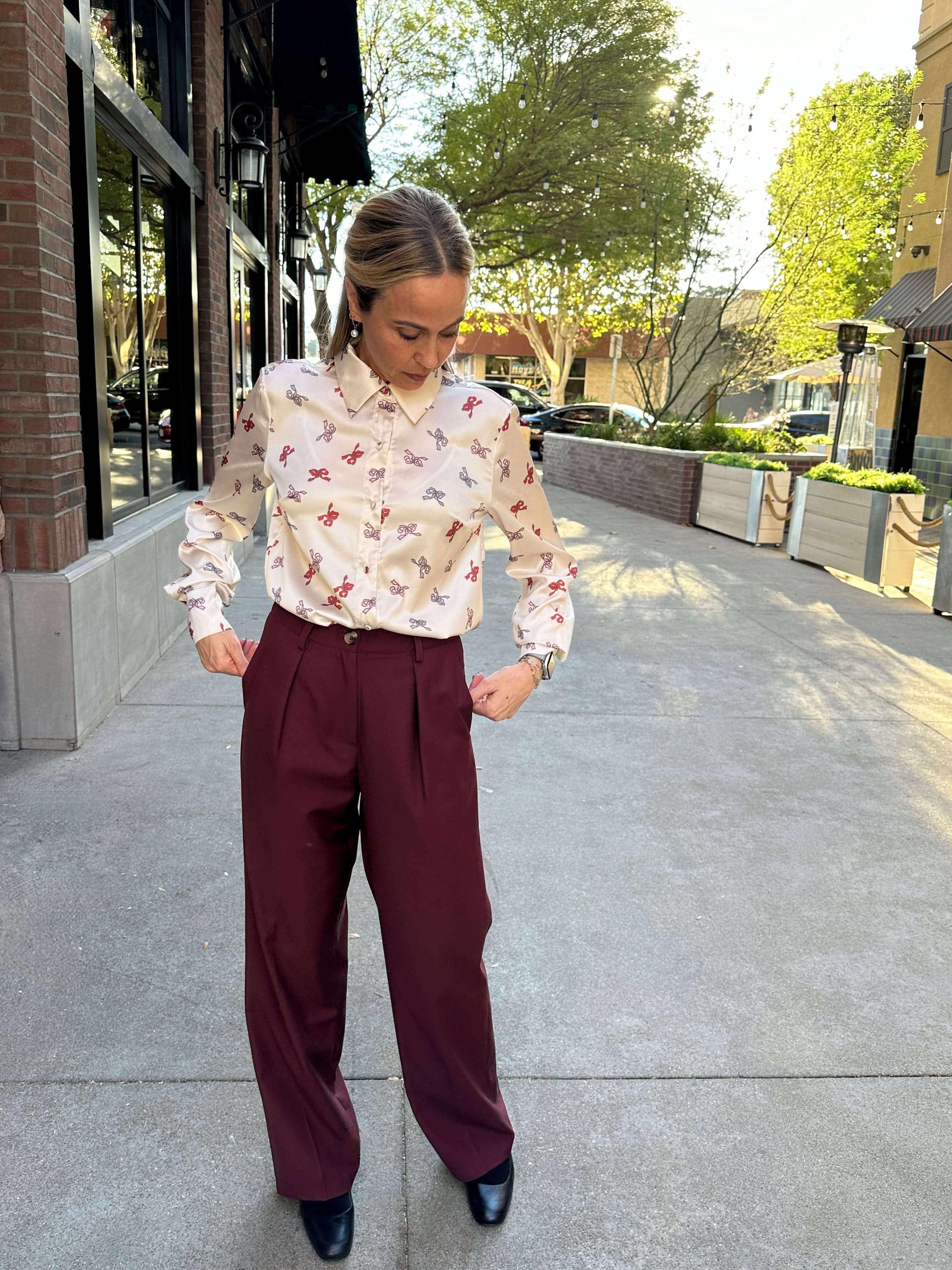 Person wearing a patterned blouse and maroon pants on a city street.