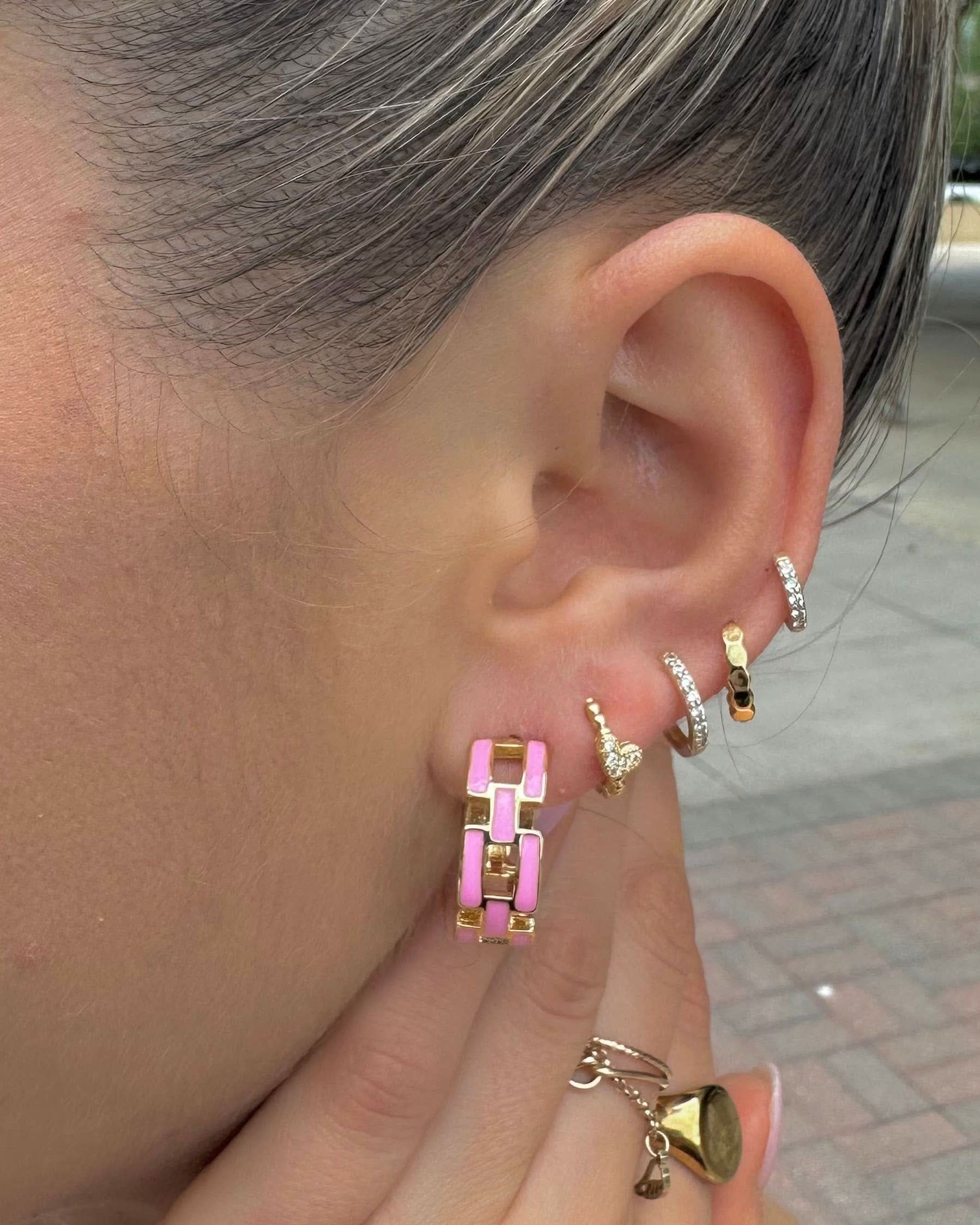 Close-up of an ear with multiple gold hoop earrings, including a pink one, with a blurred outdoor background.