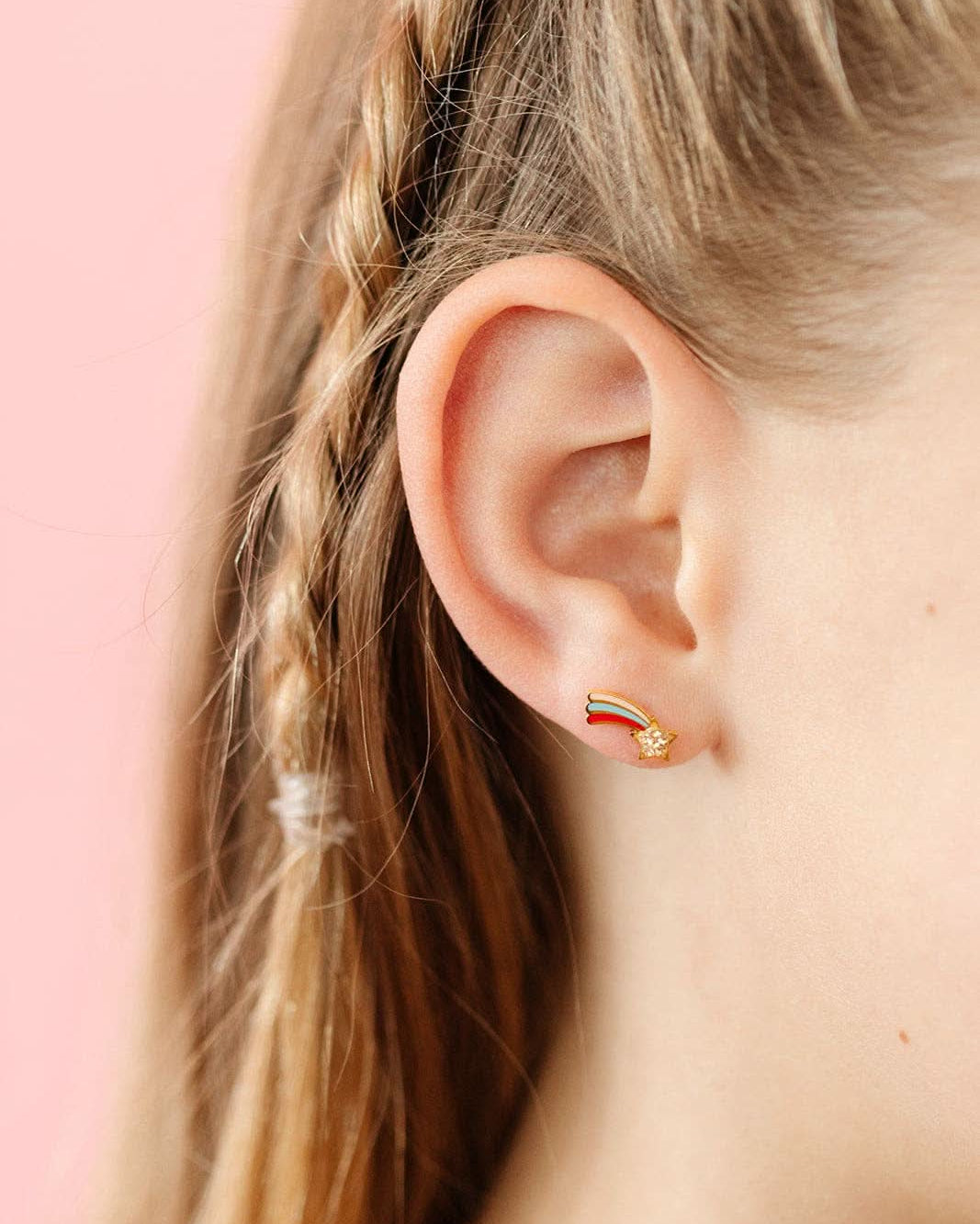 Close-up of an ear wearing a gold earring with a colorful design against a pink background