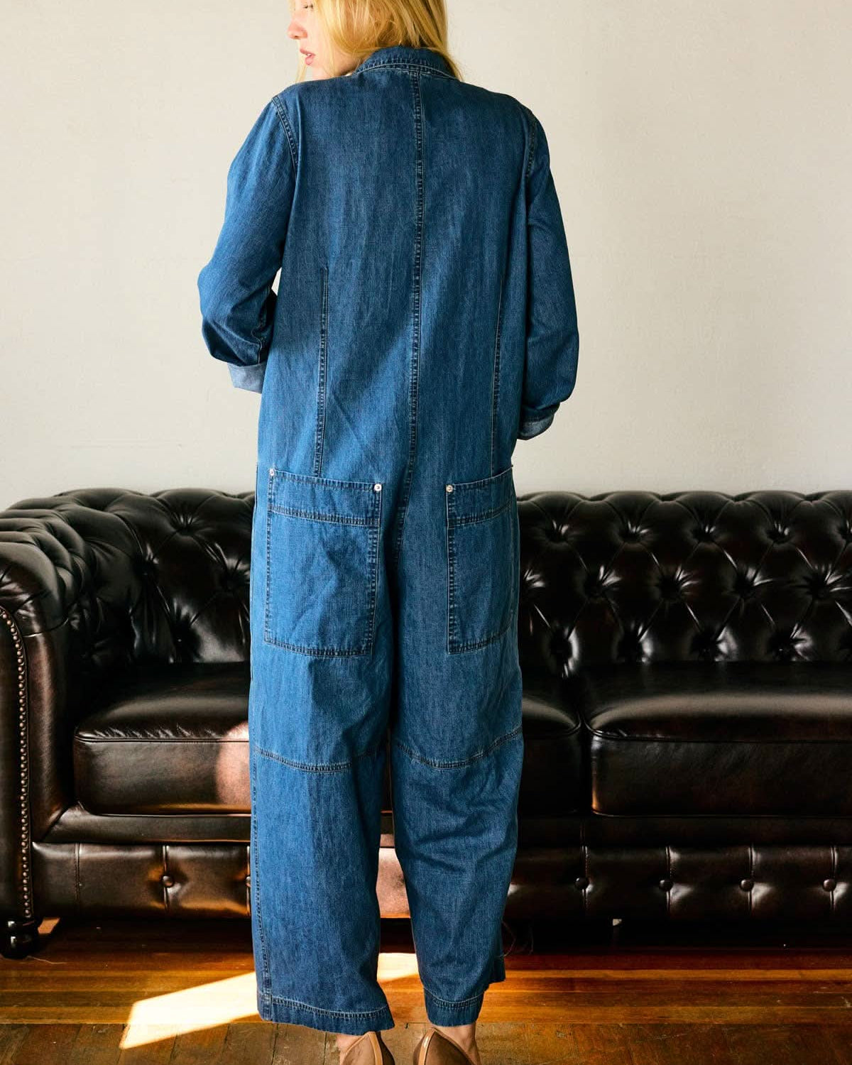 Person wearing a blue denim jumpsuit standing in front of a black leather couch.