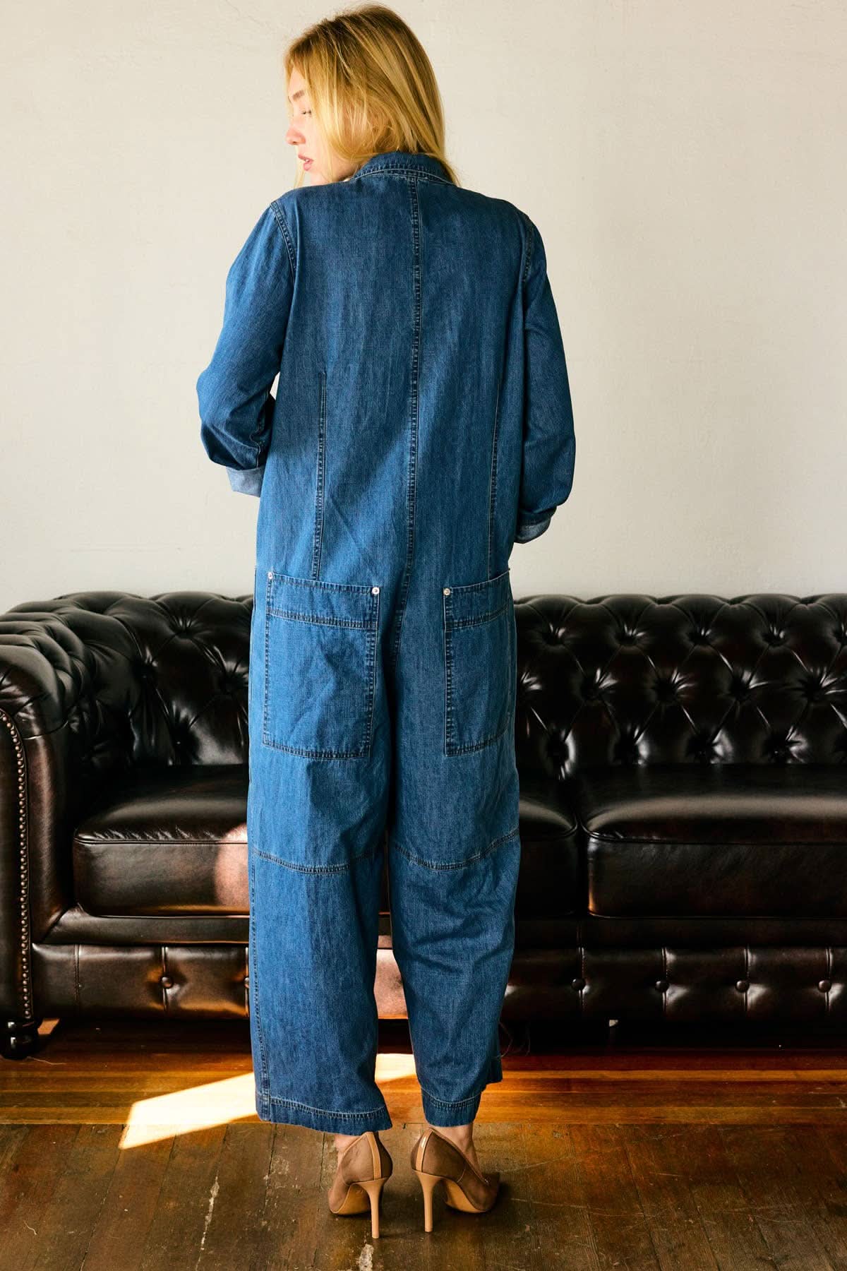 Person wearing a blue denim jumpsuit standing in front of a black leather couch.