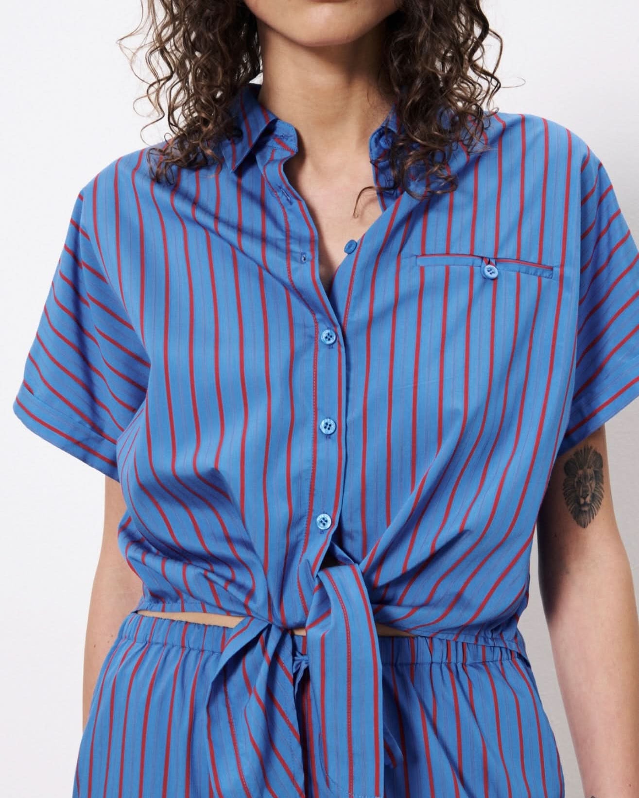 Woman wearing blue botton up blouse with vertical red stripes and a front pocket. Wearing pants with the same pattern.