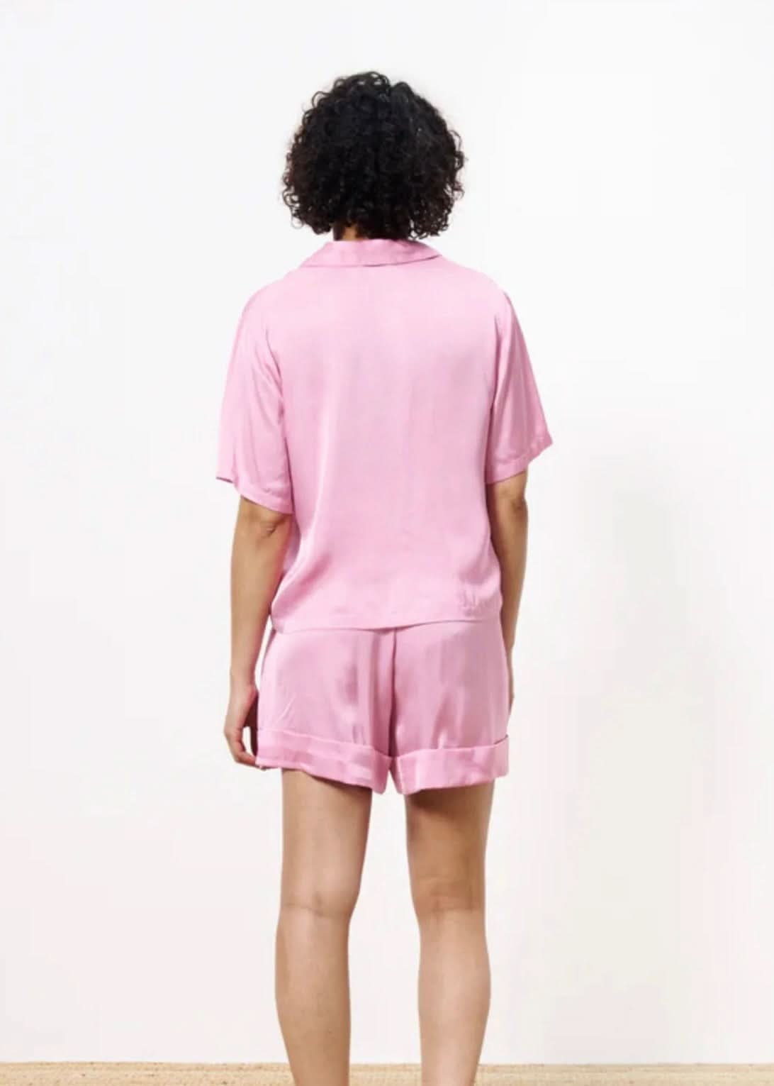 Woman turned away from the camera wearing a pink silky blouse button up with matching shorts