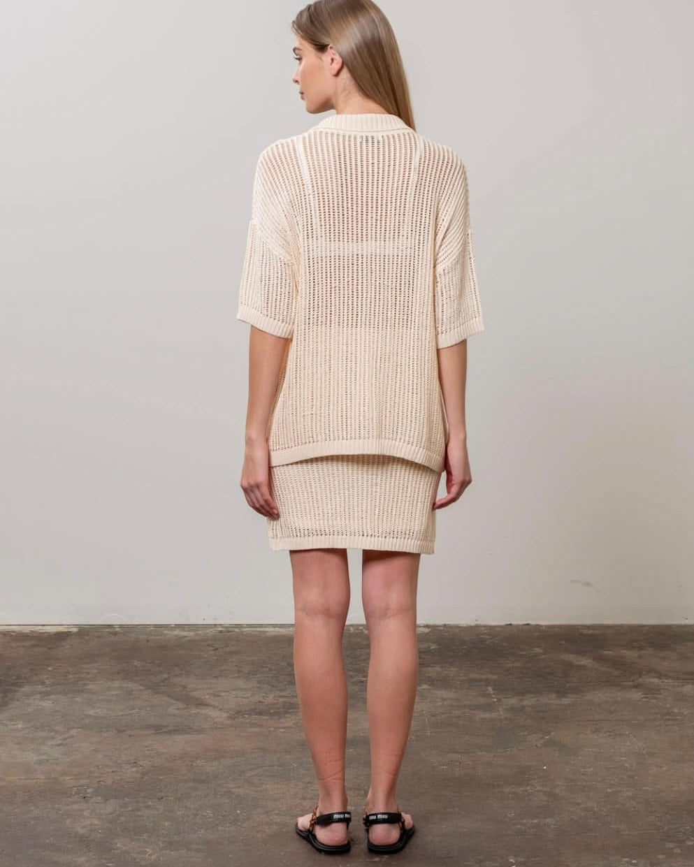Woman facing away from the camera wearing crochet tan beige colored collared button up shirt with matching skirt
