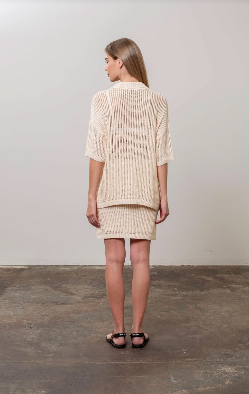 Woman facing away from the camera wearing crochet tan beige colored collared button up shirt with matching skirt