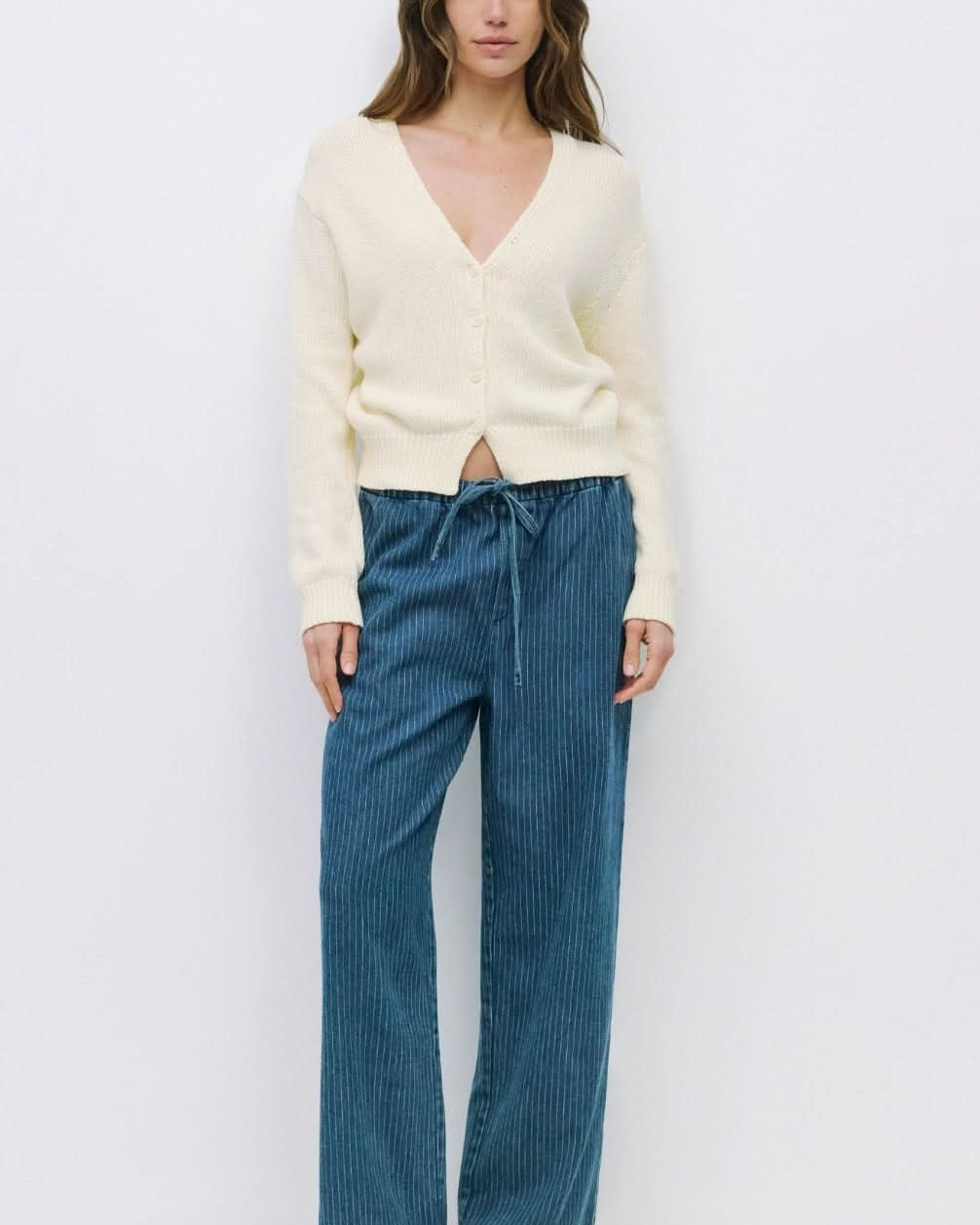 Woman wearing off white knit long sleeve sweater deep v neck with front button closure paired with pinstriped blue drawstring jeans